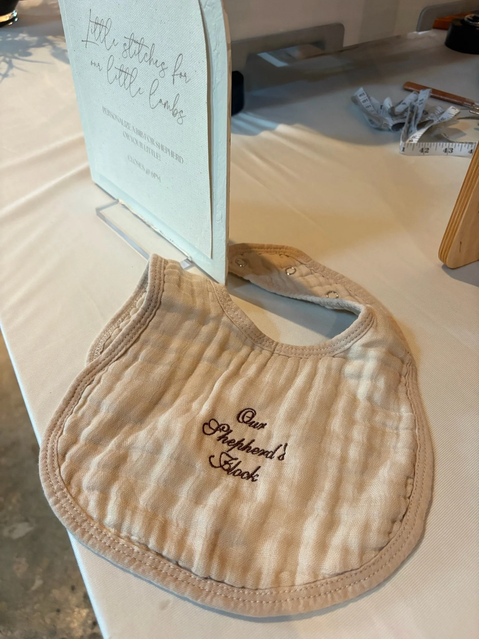 A beige quilted baby bib with embroidered text resting on a white table at a craft or retail display. In the background, there is a measuring tape and various crafting tools.
