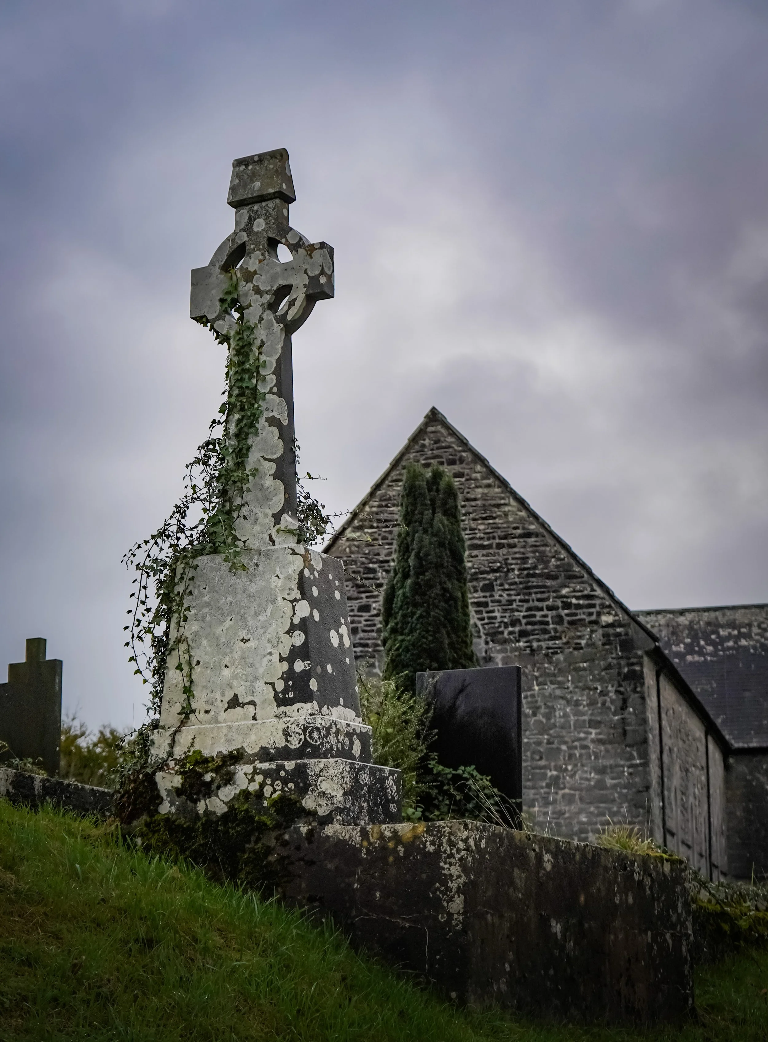 A weathered stone cross monument with climbing ivy next to a stone church building under a cloudy sky.