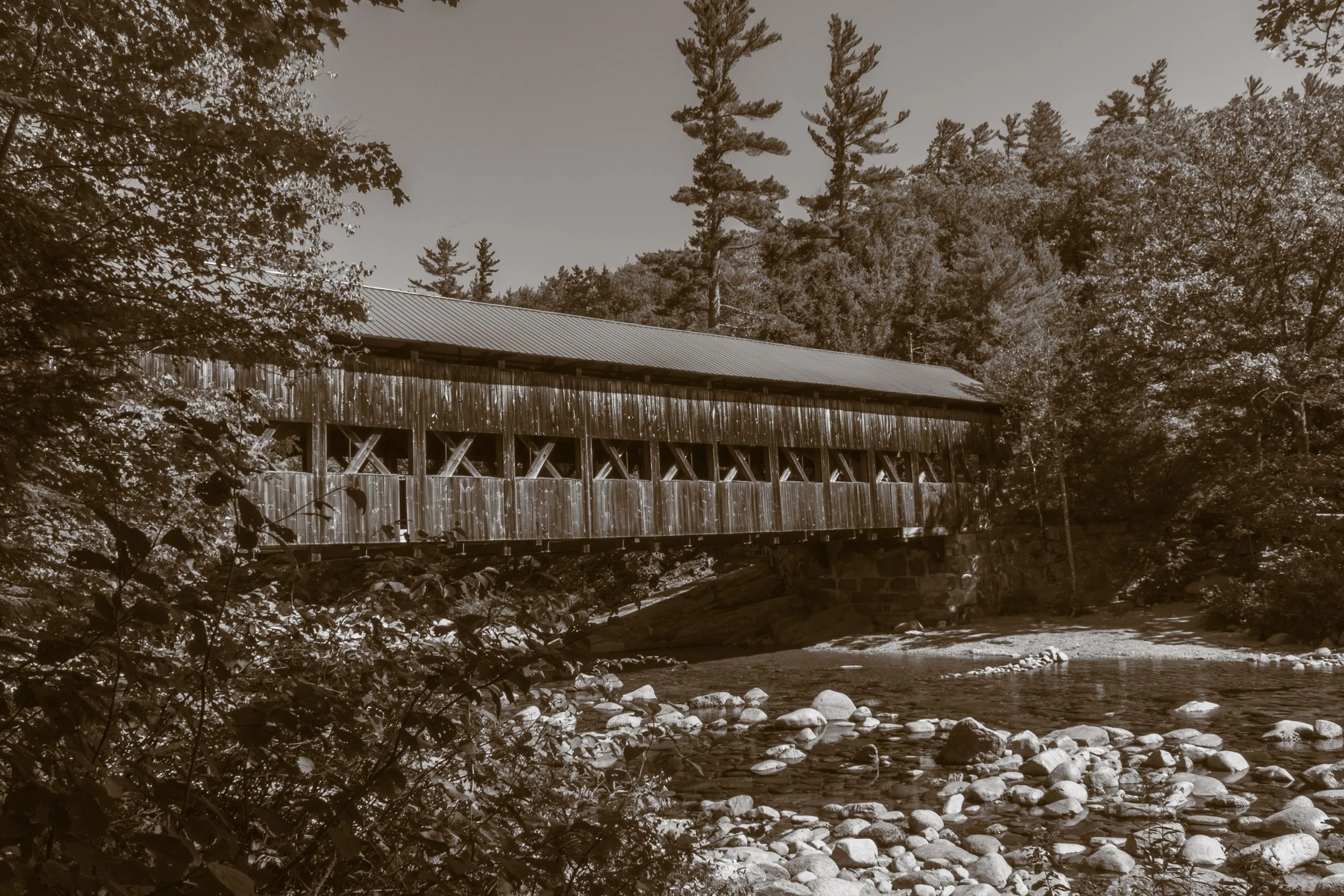 A rustic wooden covered bridge crossing over a stream in a forested area with tall trees and rocks in the water.