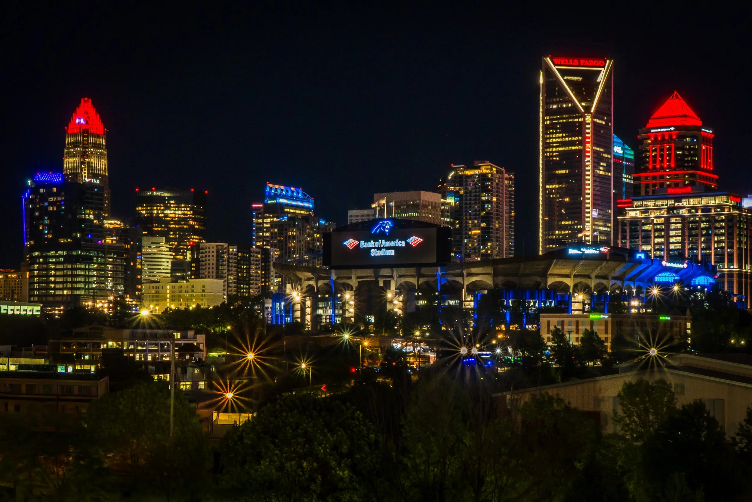 Night view of a city skyline with illuminated skyscrapers and buildings, including the Wells Fargo building, the Bank of America Stadium sign, and other colorful lights.