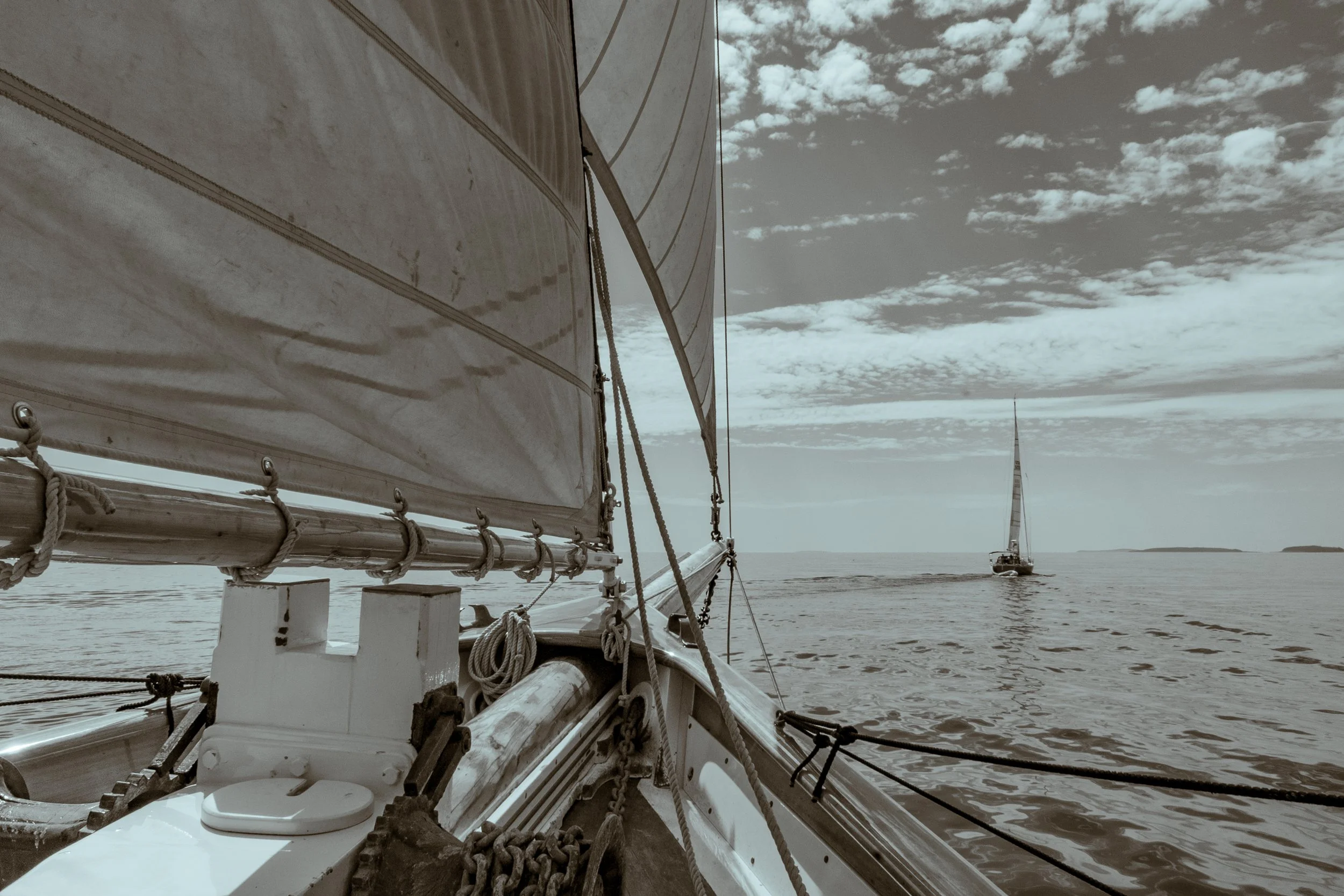 View from a sailboat showing the boat's bow, rigging, and large sail, with other sailboats in the distance on calm water under partly cloudy sky.