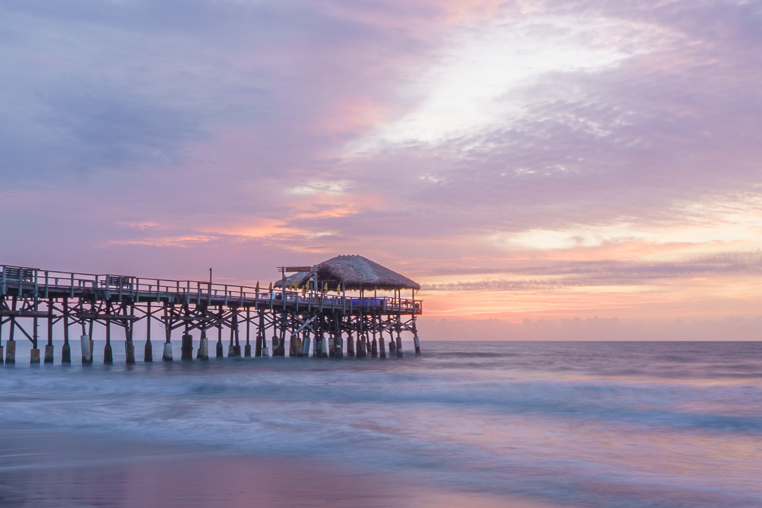 A pier extends into the ocean with a thatched roof hut at the end. The sky is painted with pastel colors of pink, purple, and orange during sunset, reflecting on the water.