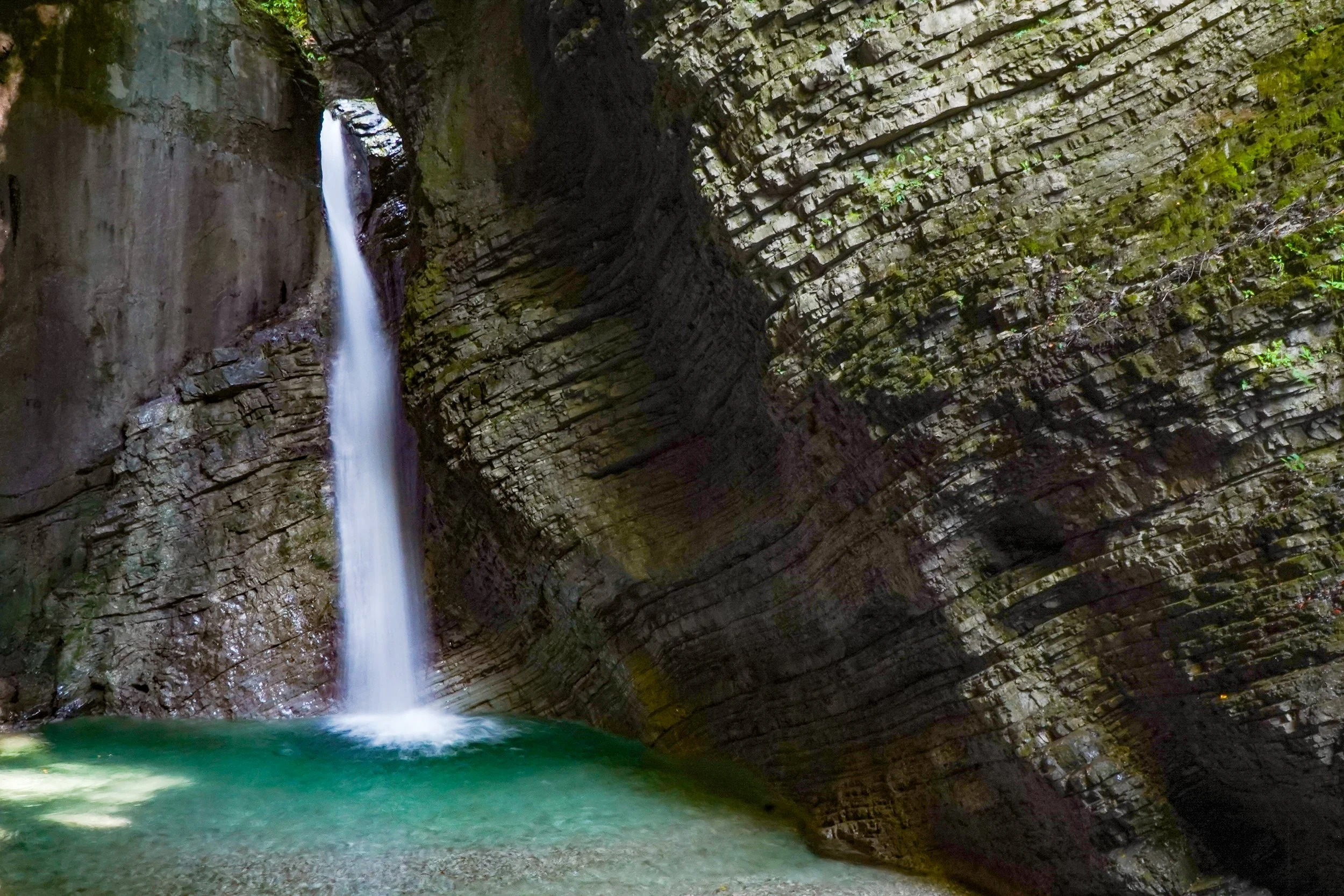 A narrow, vertically oriented cave with a waterfall flowing down into a clear pool of water at the bottom, surrounded by moss-covered rock walls.