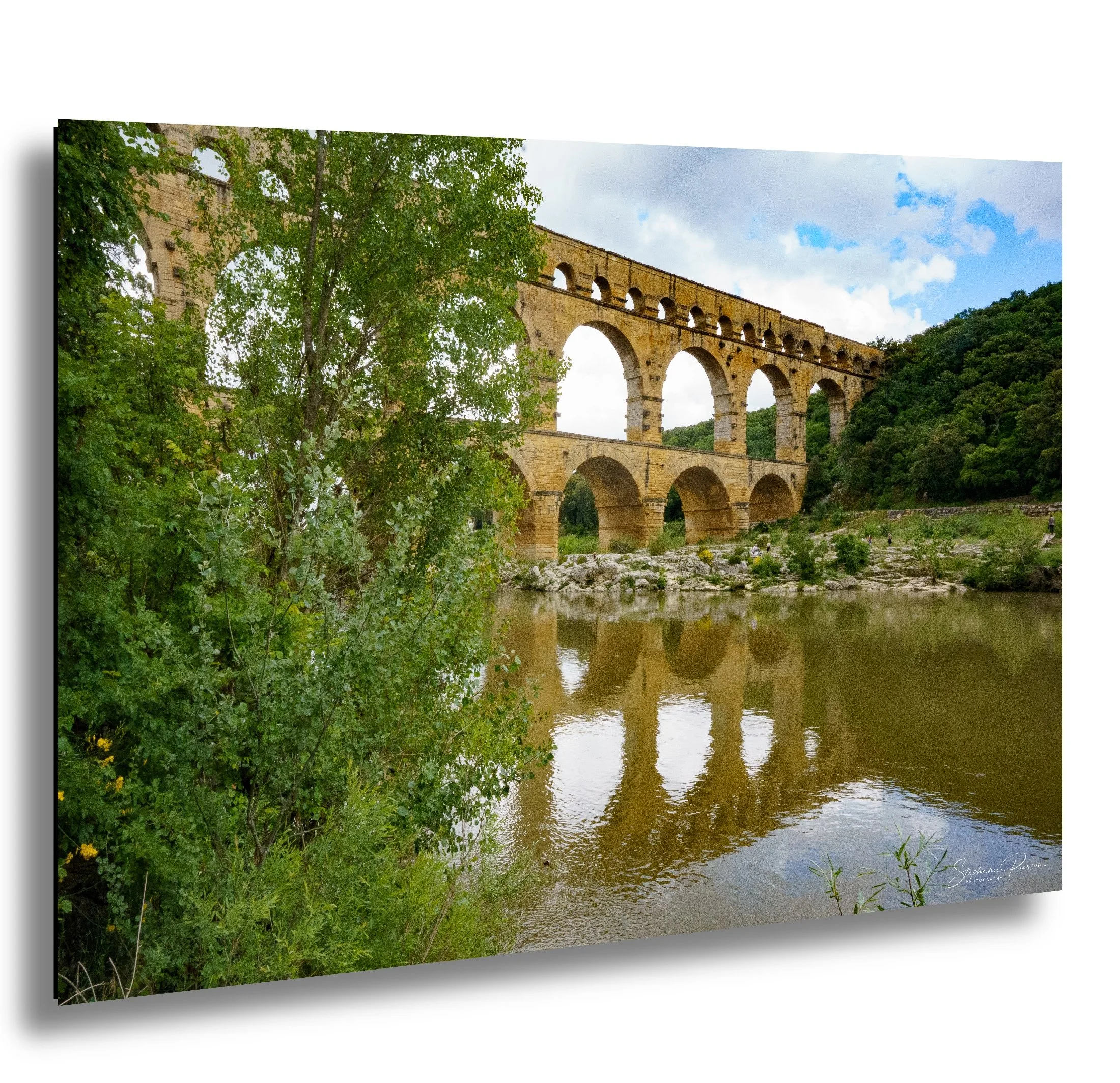 A scenic landscape featuring a historic stone aqueduct over a river, surrounded by lush green trees and a cloudy sky.