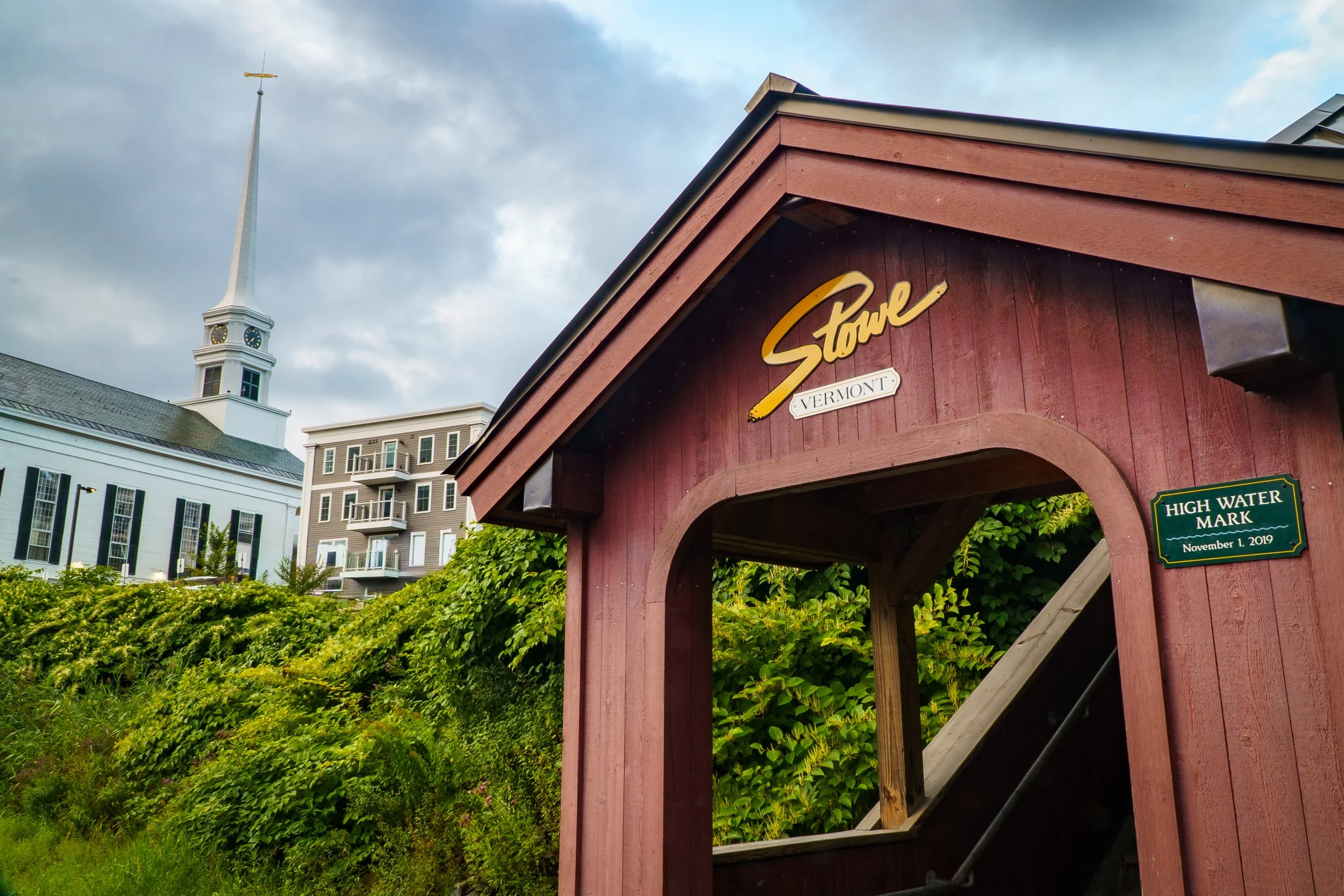 Red wooden structure with signage that reads 'Stowe Vermont' and a green plaque with 'High Water Mark November 1, 2019.' In the background, there is a white church or historic building with a tall steeple and clock, and behind it, a modern multi-stor