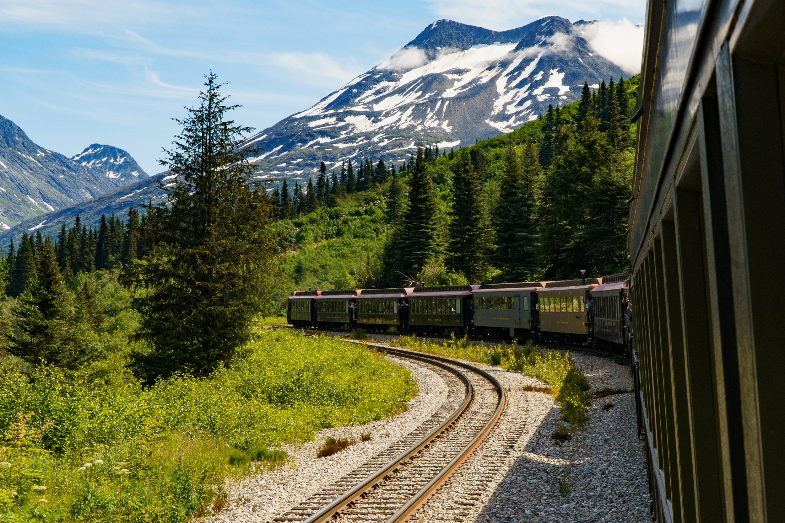 A scenic train ride through a mountainous forested area with snow-capped peaks in the background.