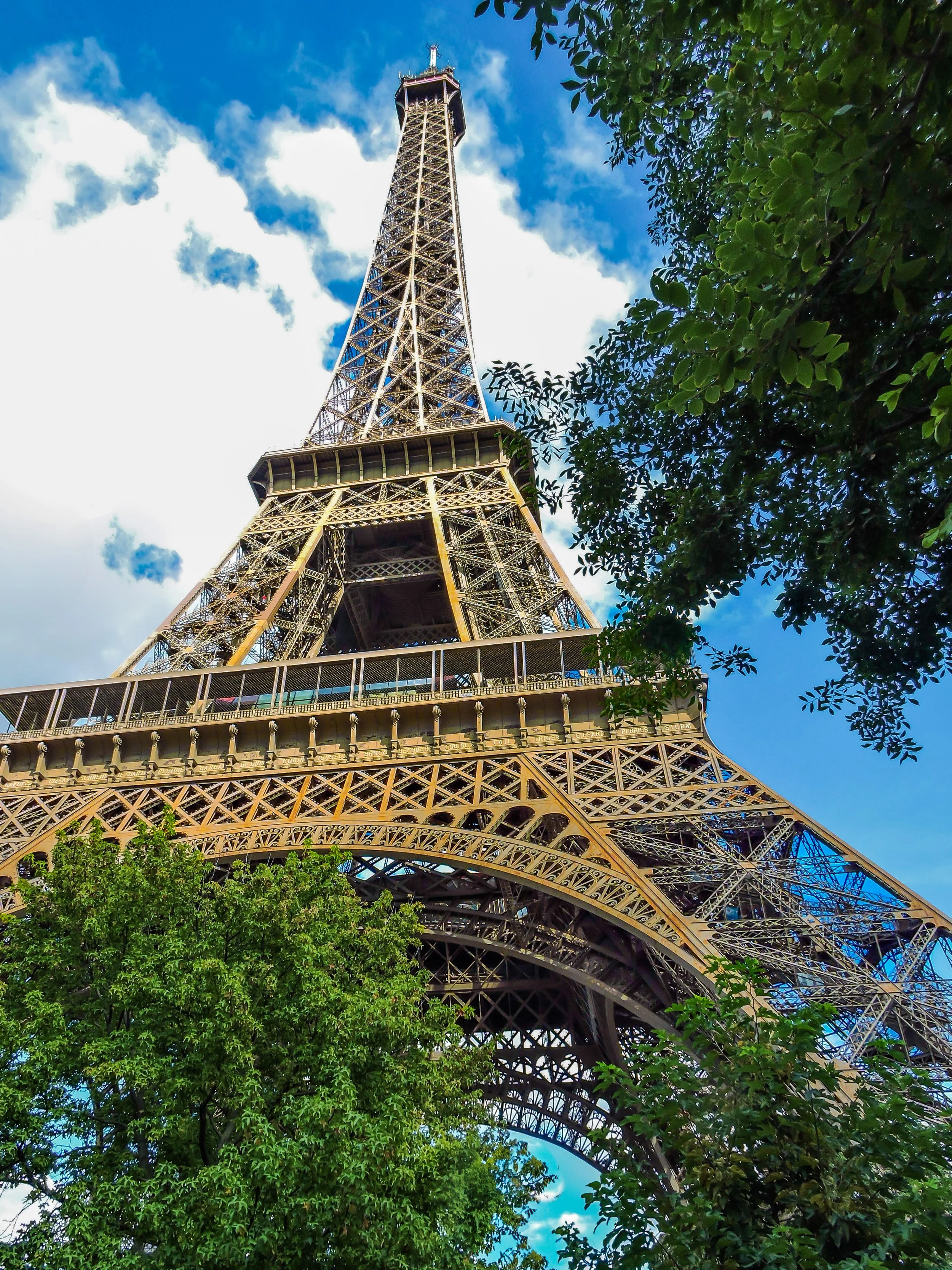 Low-angle view of the Eiffel Tower with a bright blue sky and scattered clouds, framed by green tree leaves.
