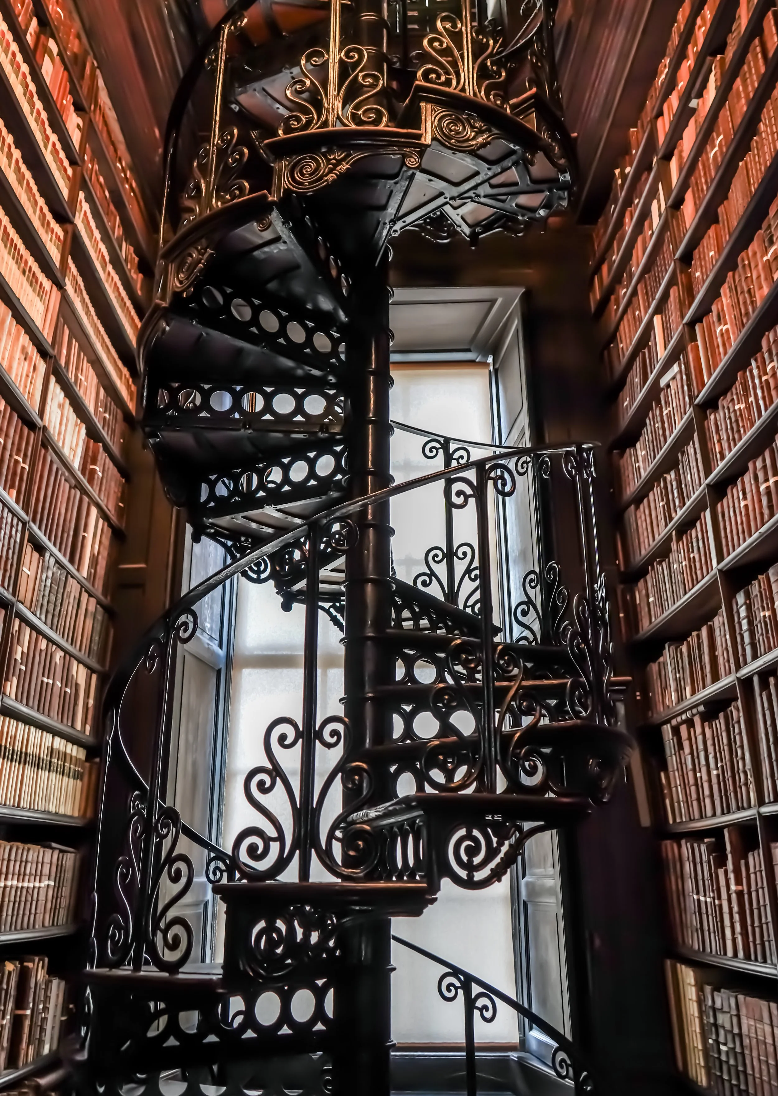 Decorative wrought iron spiral staircase in a library with floor-to-ceiling bookshelves filled with leather-bound books, sunlight coming through a window at the top of the stairs.