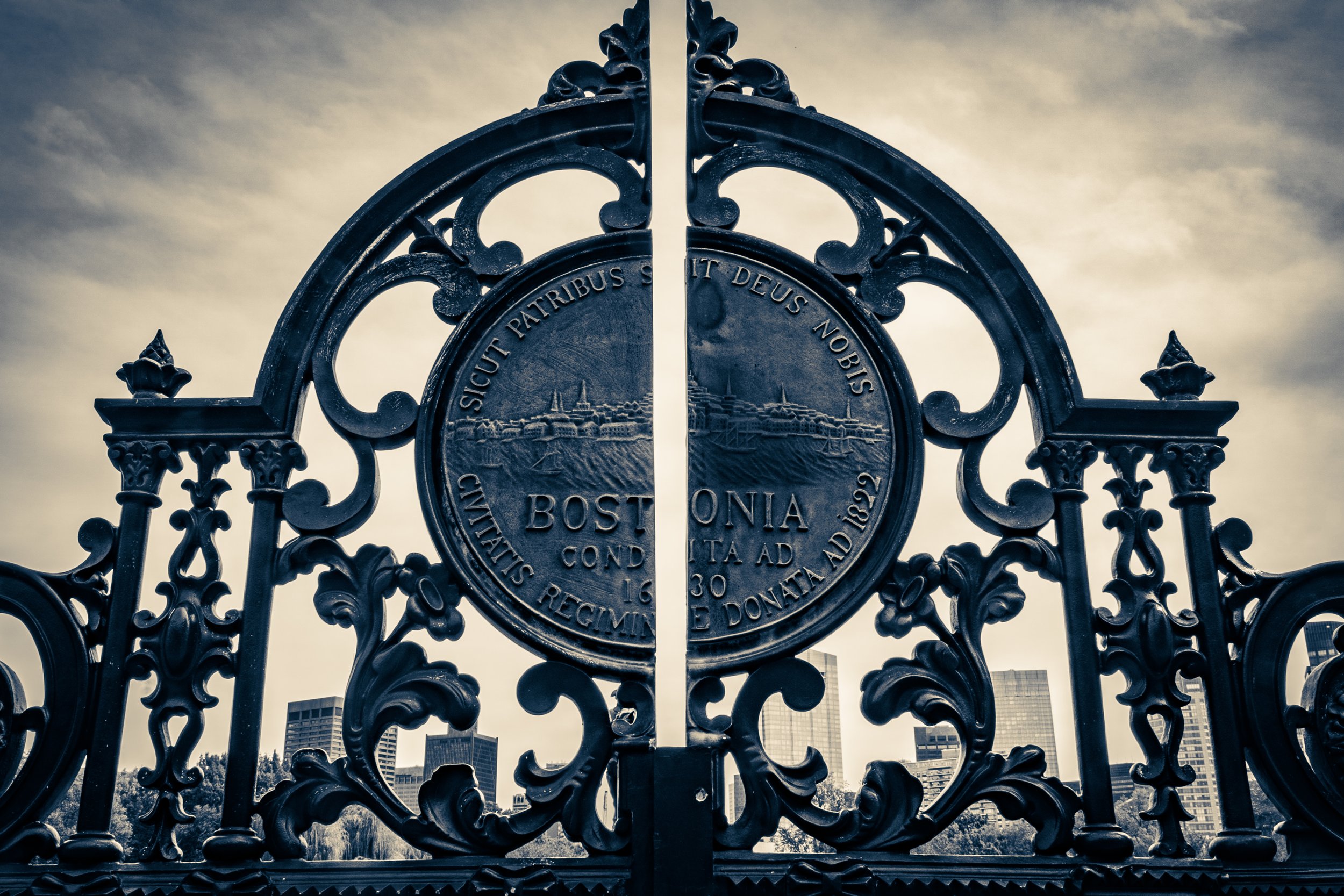 Close-up of an ornate, black wrought-iron gate with a circular emblem displaying a cityscape and Latin inscription, set against a cloudy sky and modern buildings in the background.