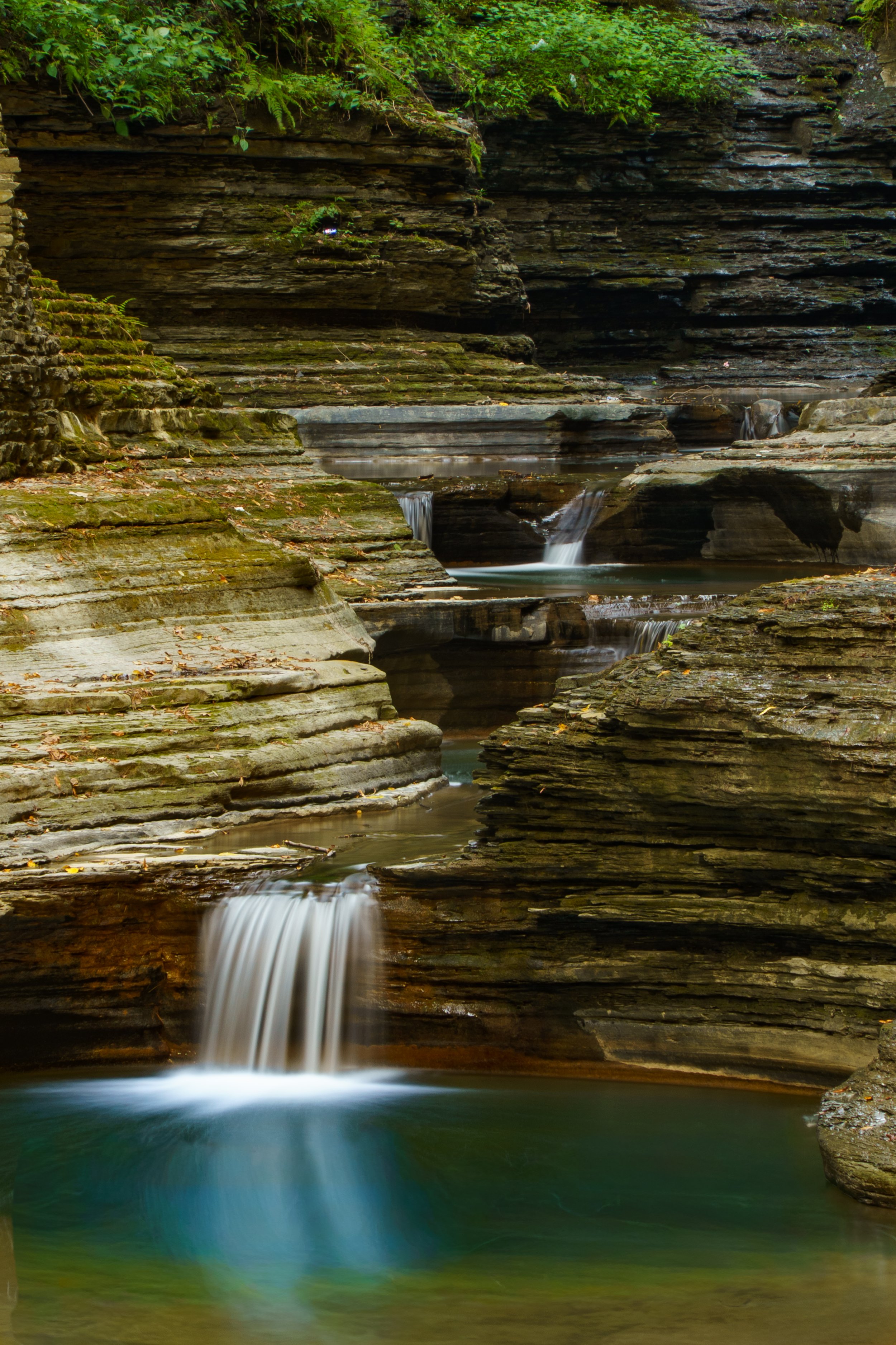 A waterfall cascading over layered rock formations in a dense green forest, with moss and foliage on the rocks and small pools of water at the base.