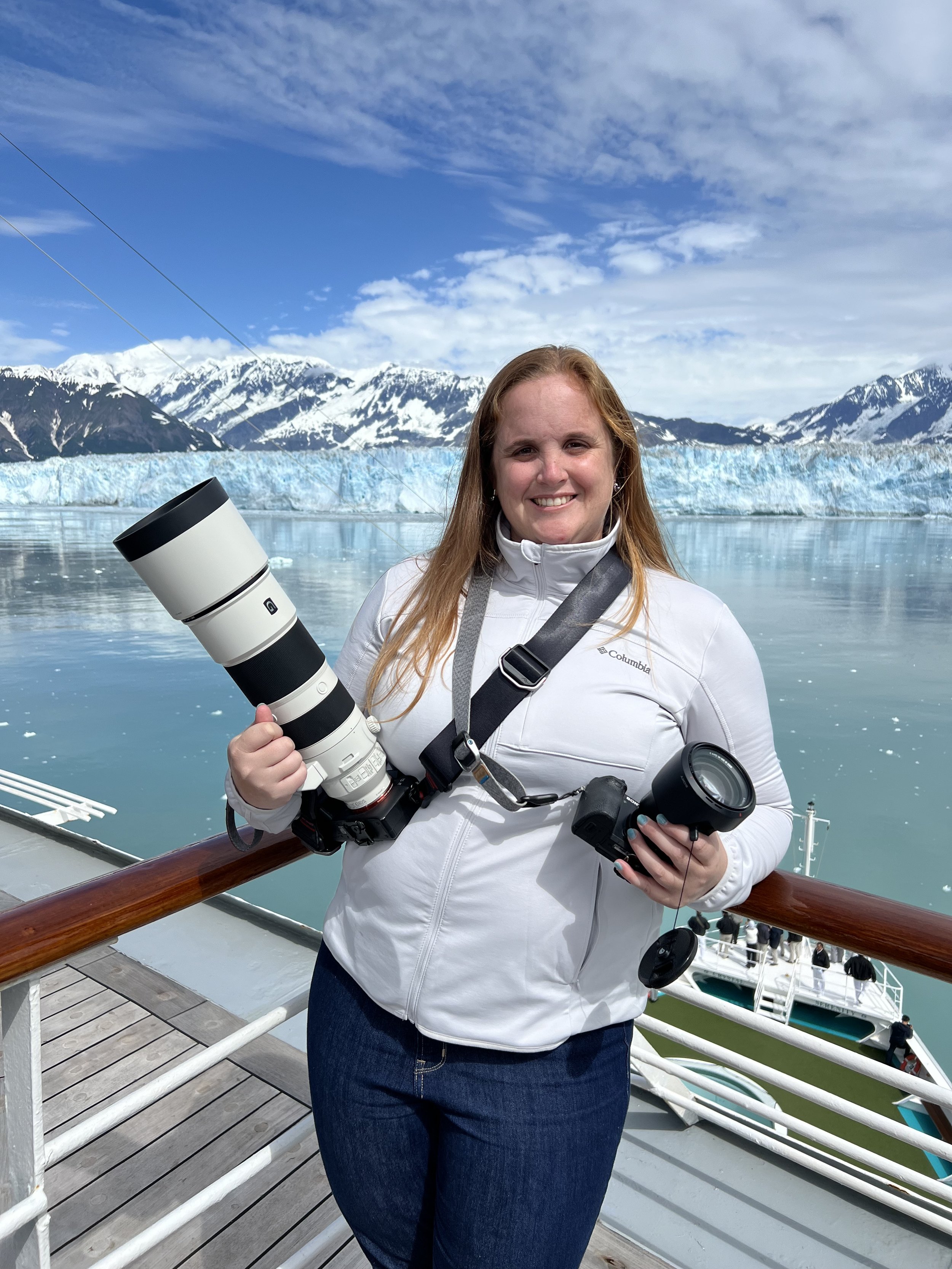 A woman standing on a boat deck with snow-capped mountains and glaciers in the background, holding a camera lens and a camera.