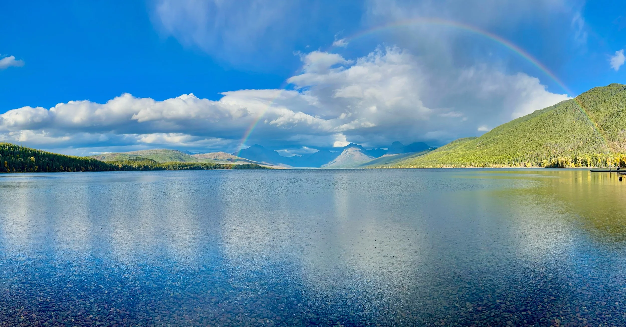 Scenic view of a large lake surrounded by green forested hills and mountains, with a rainbow arching across a partly cloudy sky.