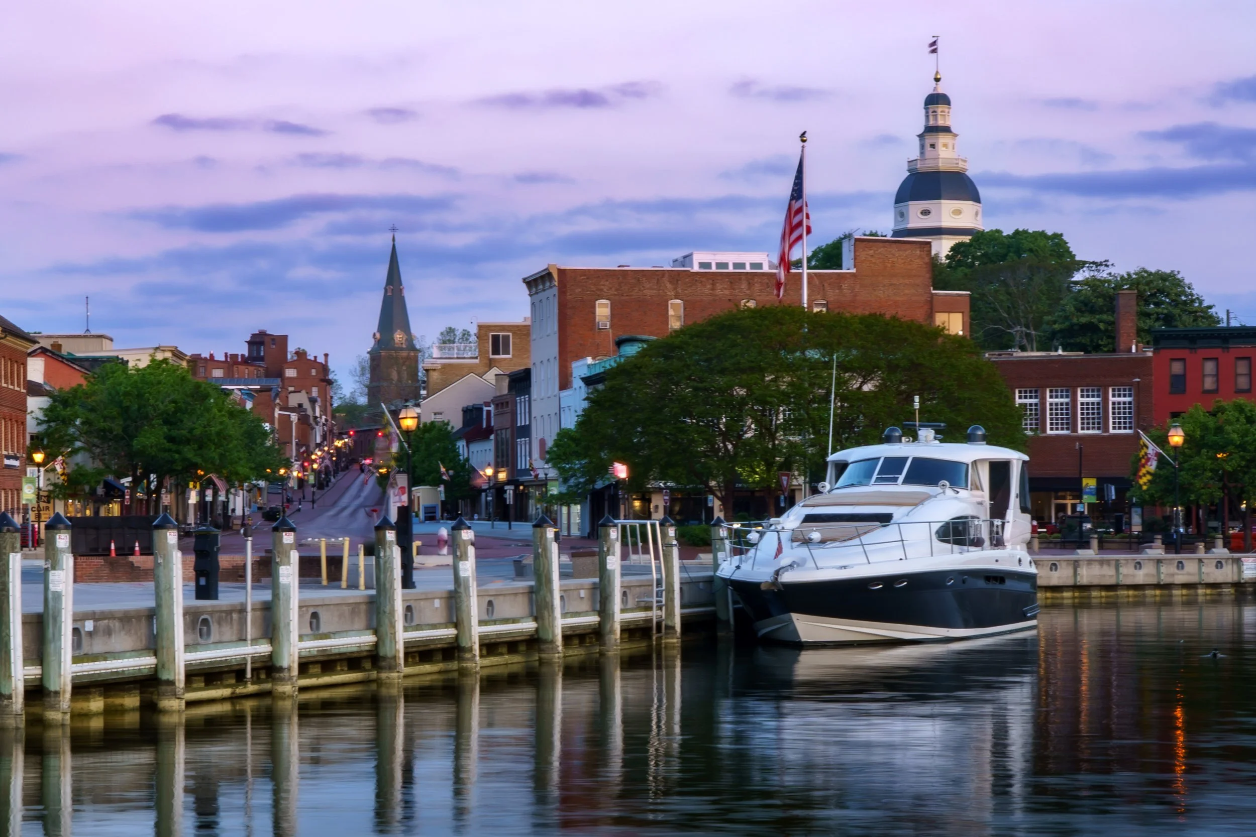 A waterfront scene with boats docked at a marina, historic brick buildings, and church steeples, with a cloudy sky overhead, in a small American town.