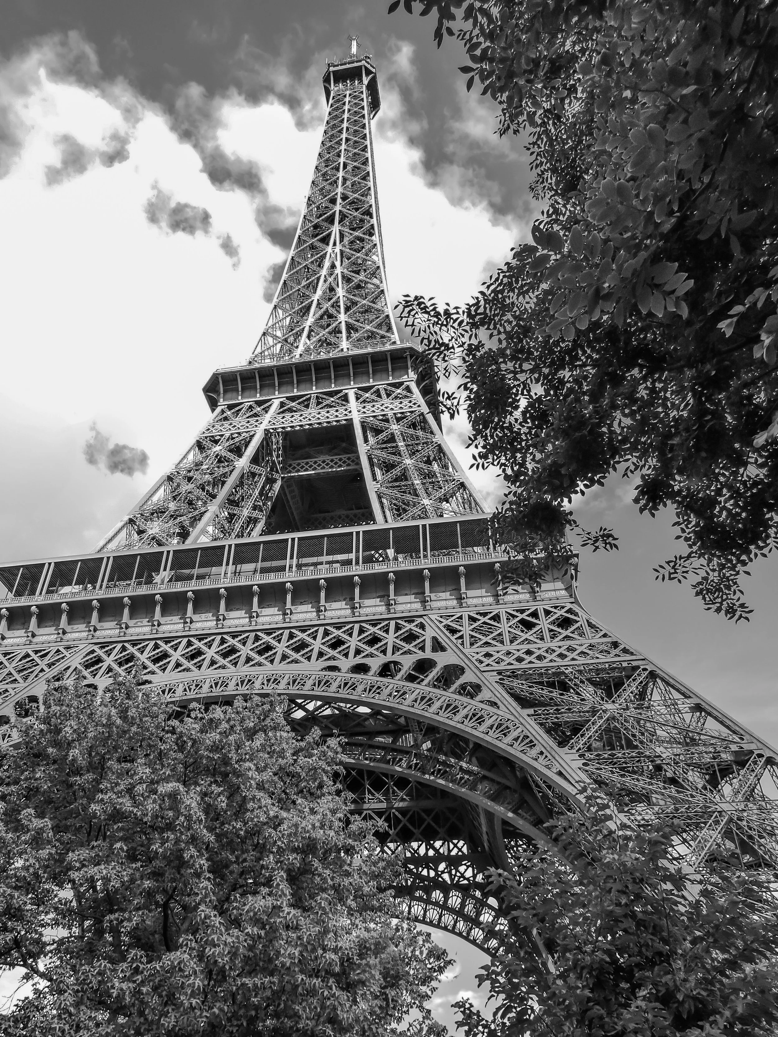 Black and white photograph of the Eiffel Tower from a low angle, partially obscured by trees and clouds in the sky.