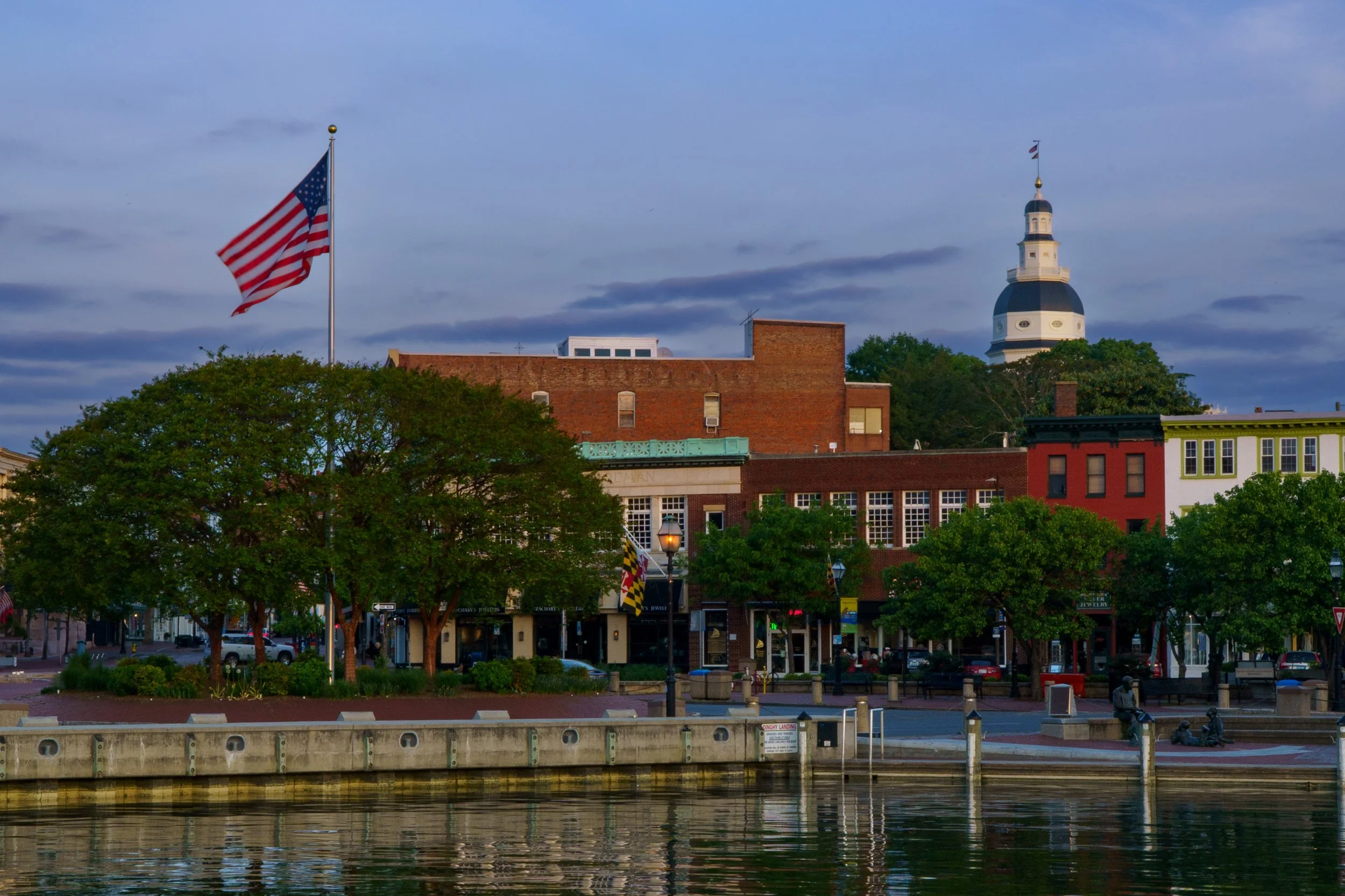 A town scene with buildings, trees, and streetlamp along a waterfront. An American flag is flying on a tall flagpole, with a historic-looking church with a domed steeple in the background, under a cloudy sky.