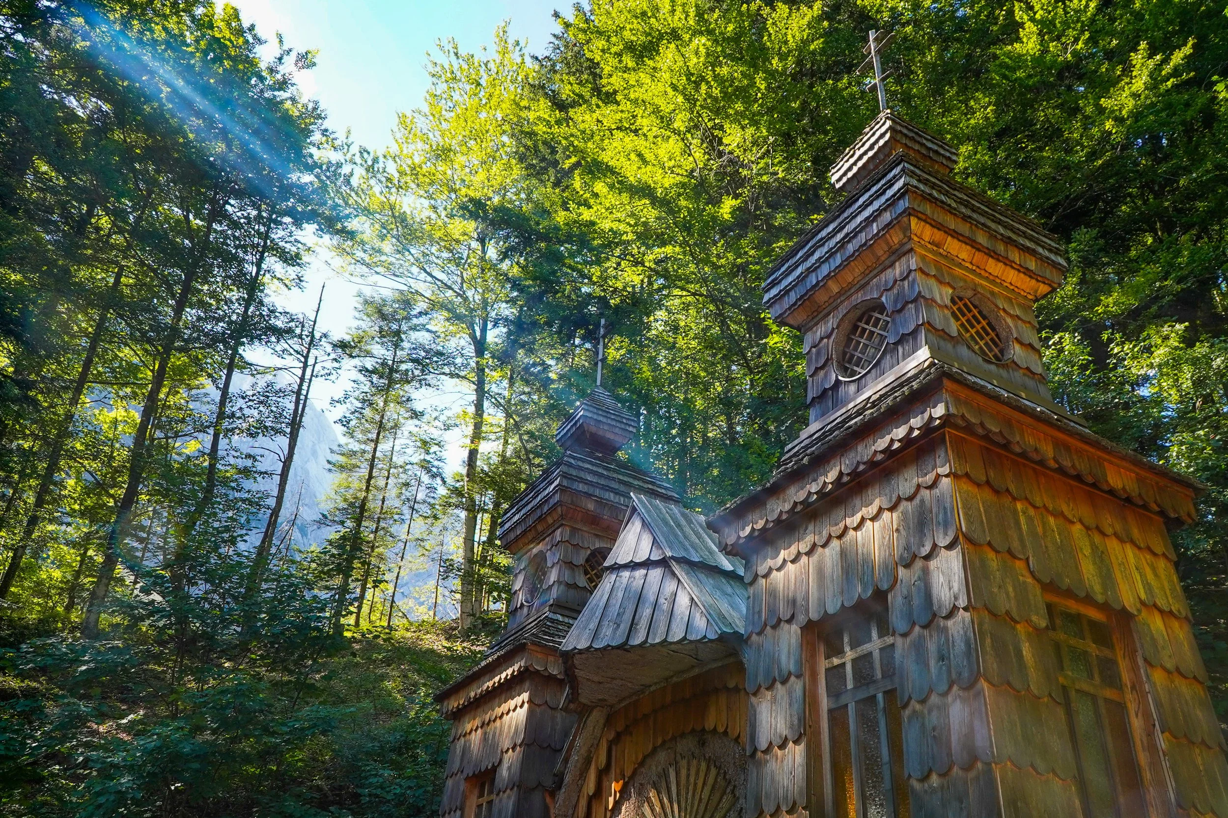A wooden church with multiple steeples nestled among tall green trees, with sunlight filtering through the leaves.
