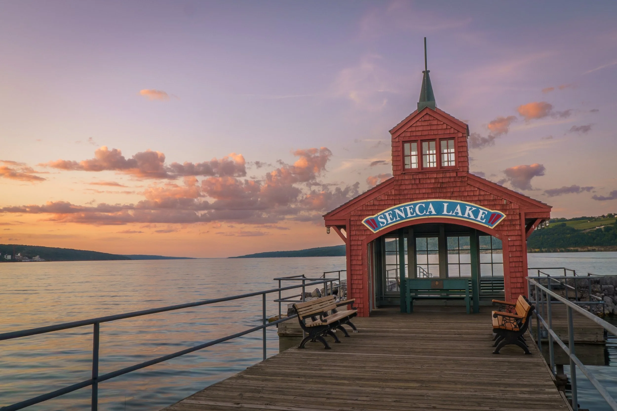Sunset view of Seneca Lake with a red wooden pavilion on a dock, benches, and a calm lake under a colorful sky with scattered clouds.