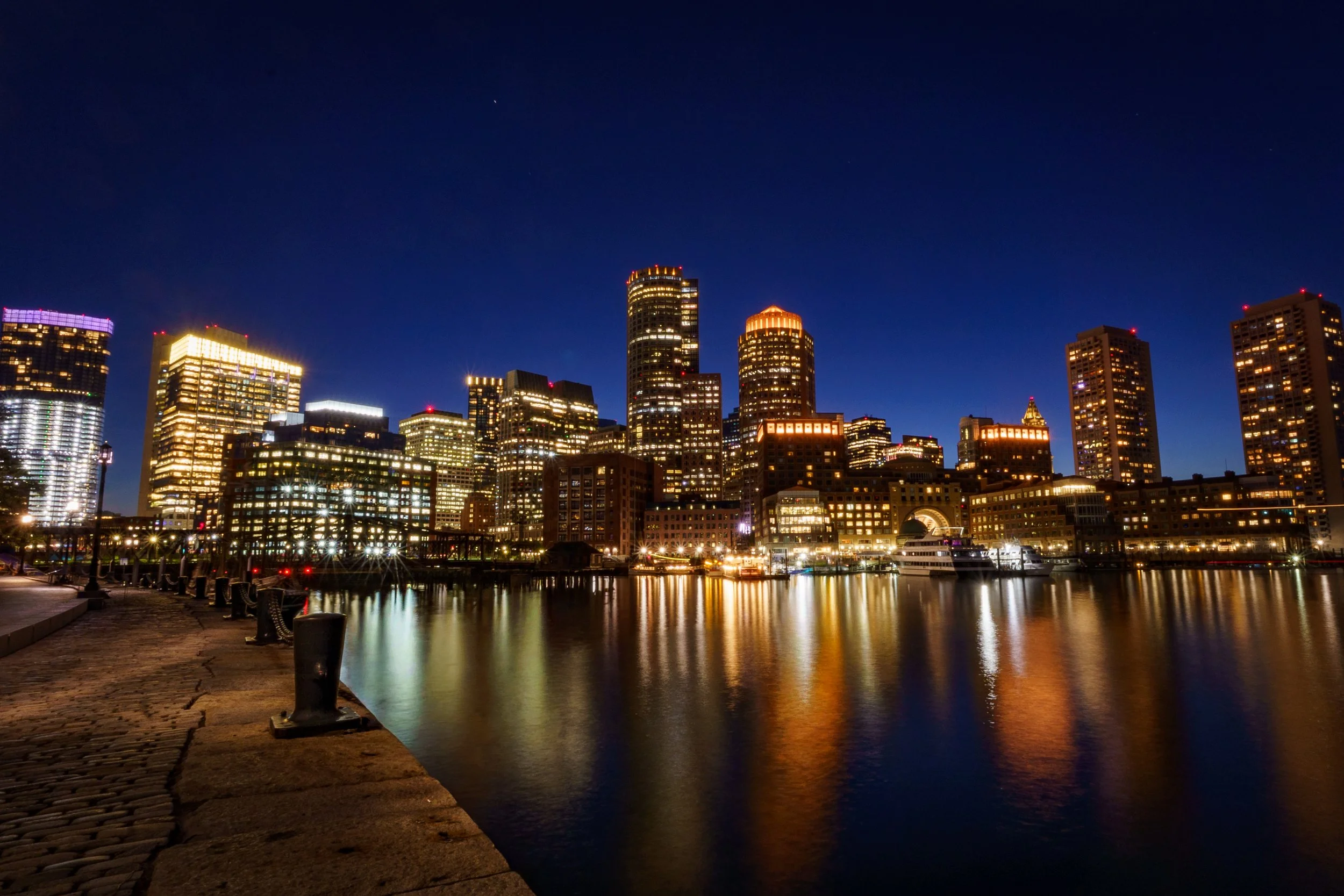 Night view of a city skyline with illuminated skyscrapers and their reflections on a calm river, with buildings lit up in various colors, and a clear dark blue sky.