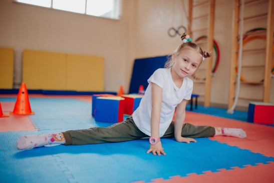 Young girl performing a split stretch on blue and red gym mats in a playroom.