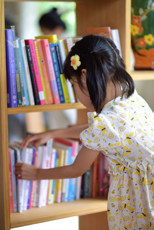 A young girl with a yellow flower hair clip browsing books on a wooden bookshelf.