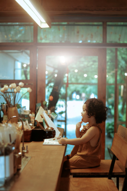 A young girl sitting at a counter in a cafe, using a touchscreen device, with sunlight shining through large windows behind her.