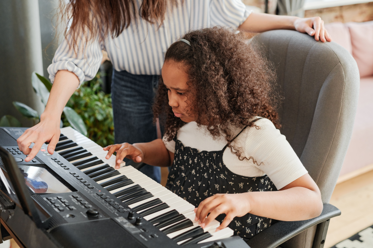 A young girl with curly hair sitting at a digital piano, playing with focus, while an adult guides her by pointing at the keys.