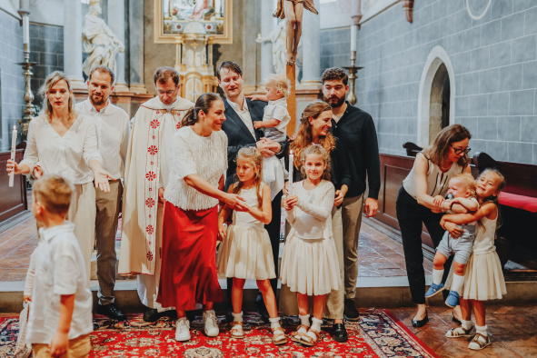 Family and children gathered inside a church for a religious ceremony, standing on a decorative red rug with an altar and religious statues in the background.
