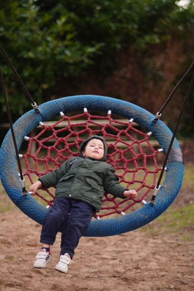 Child lying on a red netted swing with a blue circular frame, outdoors in a grassy area with trees in the background.