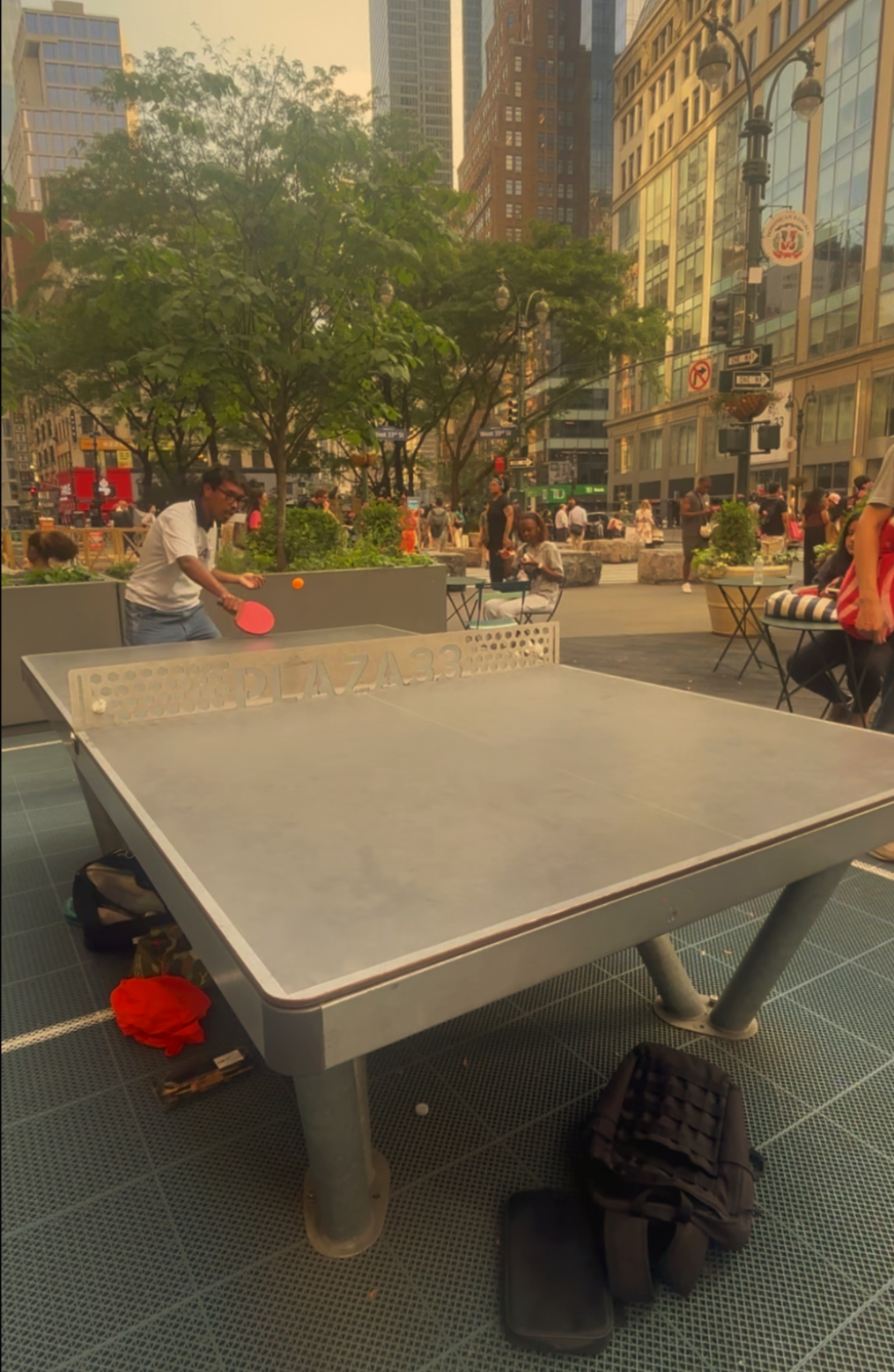 People playing ping pong outdoors in an urban plaza with tall buildings, trees, and streetlights in the background during daytime.