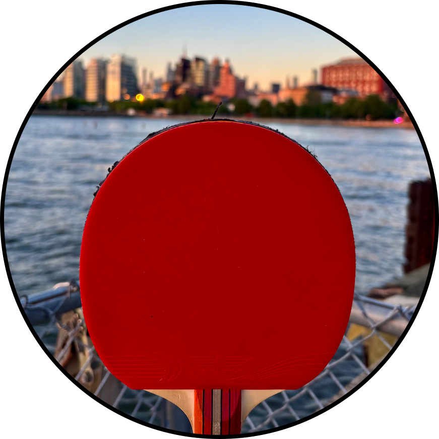A red ping pong paddle held in front of a river with a city skyline in the background during sunset.