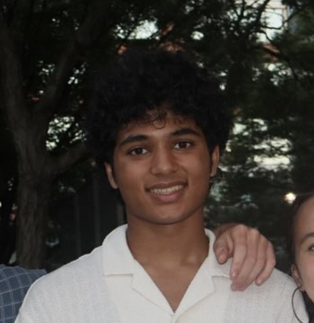Young man with curly black hair smiling, wearing a white polo shirt, outdoors with trees in the background.