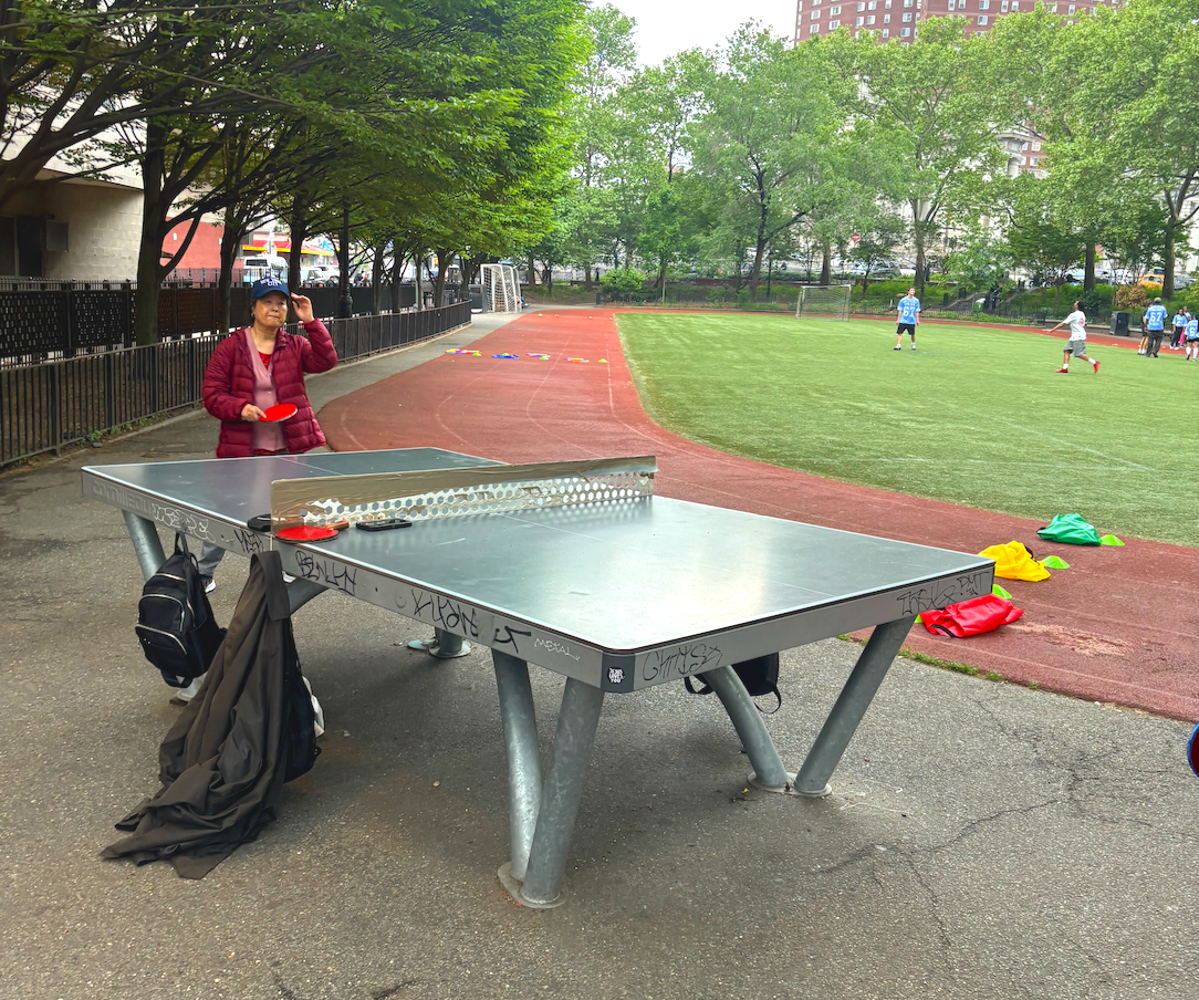 A woman standing next to an outdoor ping pong table on a playground, with a grassy athletic field and kids playing in the background. Trees line the area, and backpacks and clothing are on the ground near the table.