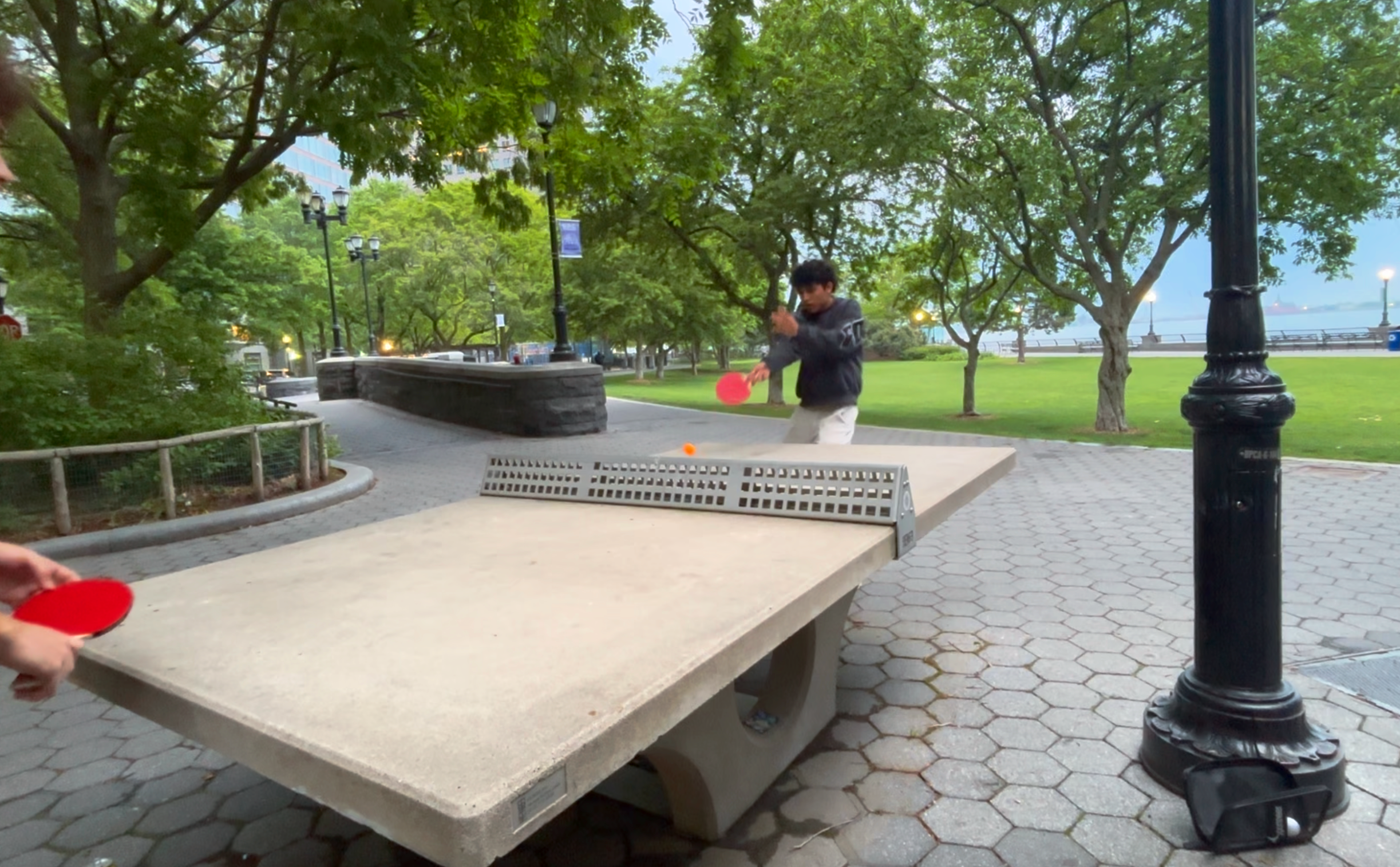 A person in a dark hoodie playing ping pong outdoors in a park with green trees, street lamps, and a waterfront in the background. Another person's hand holding a red paddle is visible in the foreground.