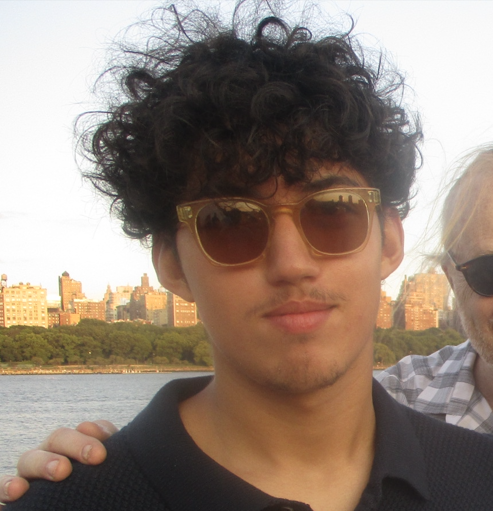 A young man with curly dark hair wearing sunglasses, standing outdoors near a body of water with a city skyline in the background.
