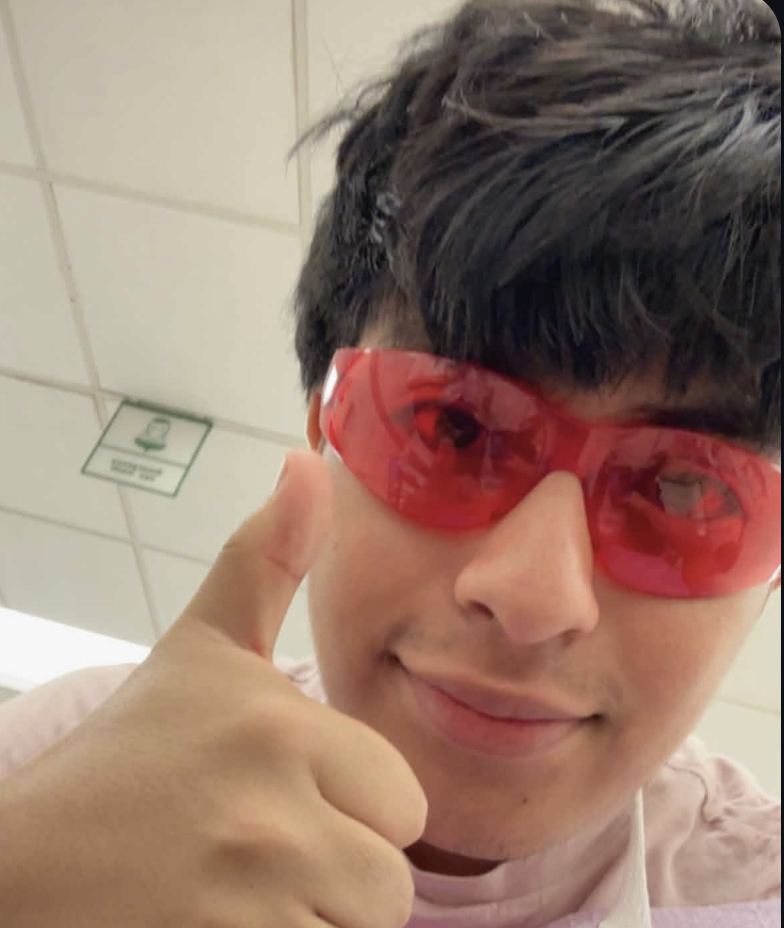 A young person wearing red sunglasses, giving a thumbs-up, indoors, with a ceiling and a sign on the ceiling visible in the background.