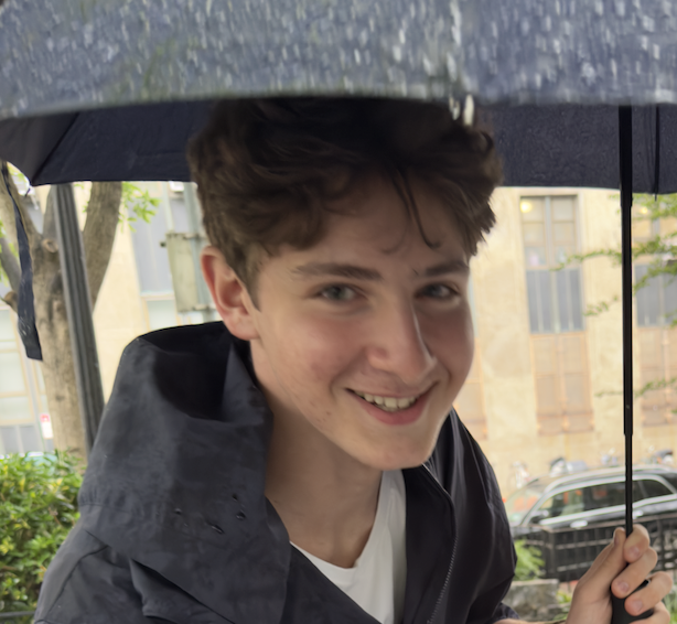 A smiling young man holding an umbrella in rainy weather outside.
