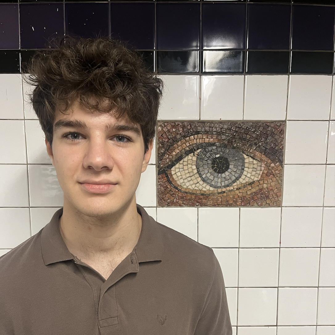 A young man with curly brown hair and fair skin standing in front of a wall of white square tiles. Behind him, the tiles include a mosaic of a human eye with a black pupil, gray iris, and brown and white sclera, set in a mosaic framed by dark and reddish tiles.