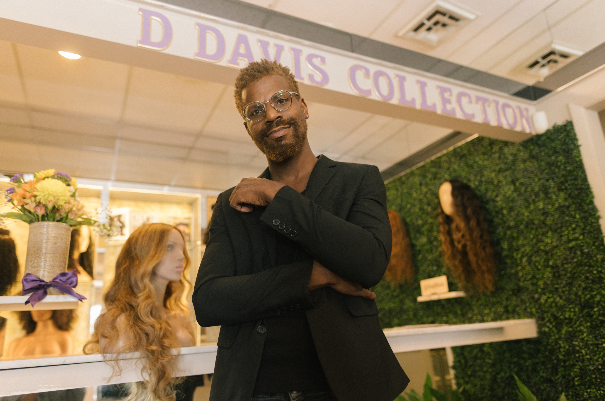 A man with glasses and a beard in a black blazer posing with his arms crossed, standing inside a store or exhibit called 'Daisy's Collection,' with mannequins of hairless and curly-haired dolls or mannequins in the background.