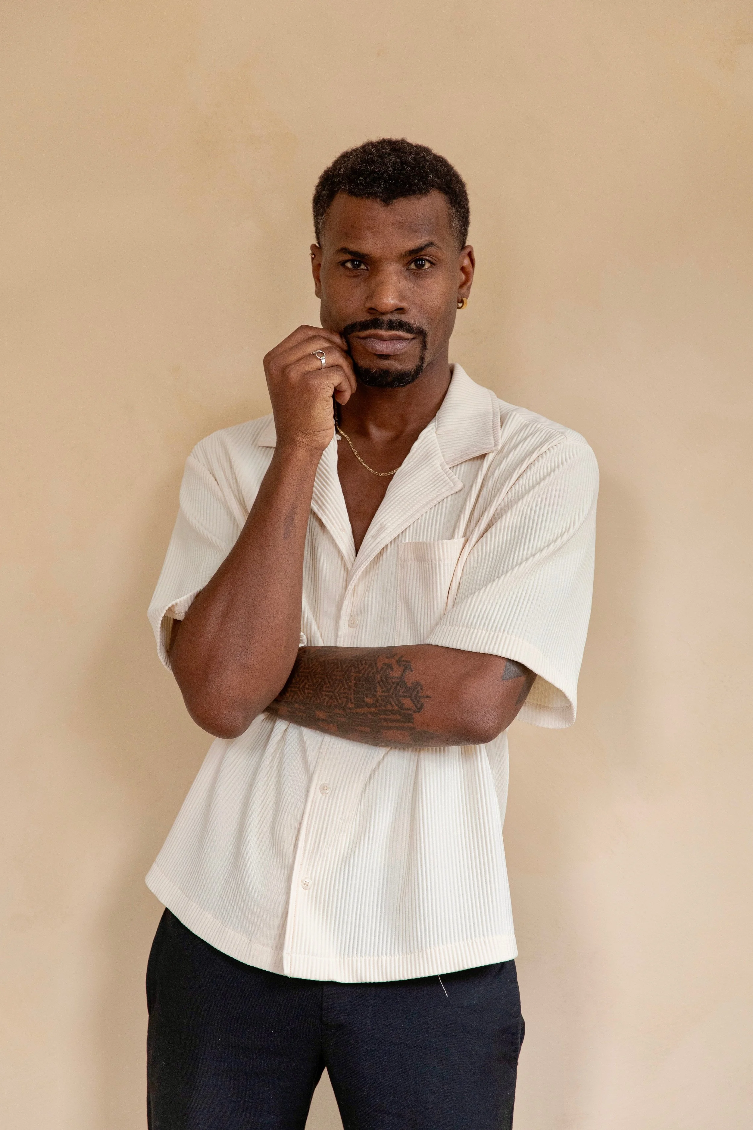 A man wearing a short-sleeved cream-colored shirt with black pants, standing against a plain beige wall, with one hand touching his face and the other crossed over his chest, looking directly at the camera.