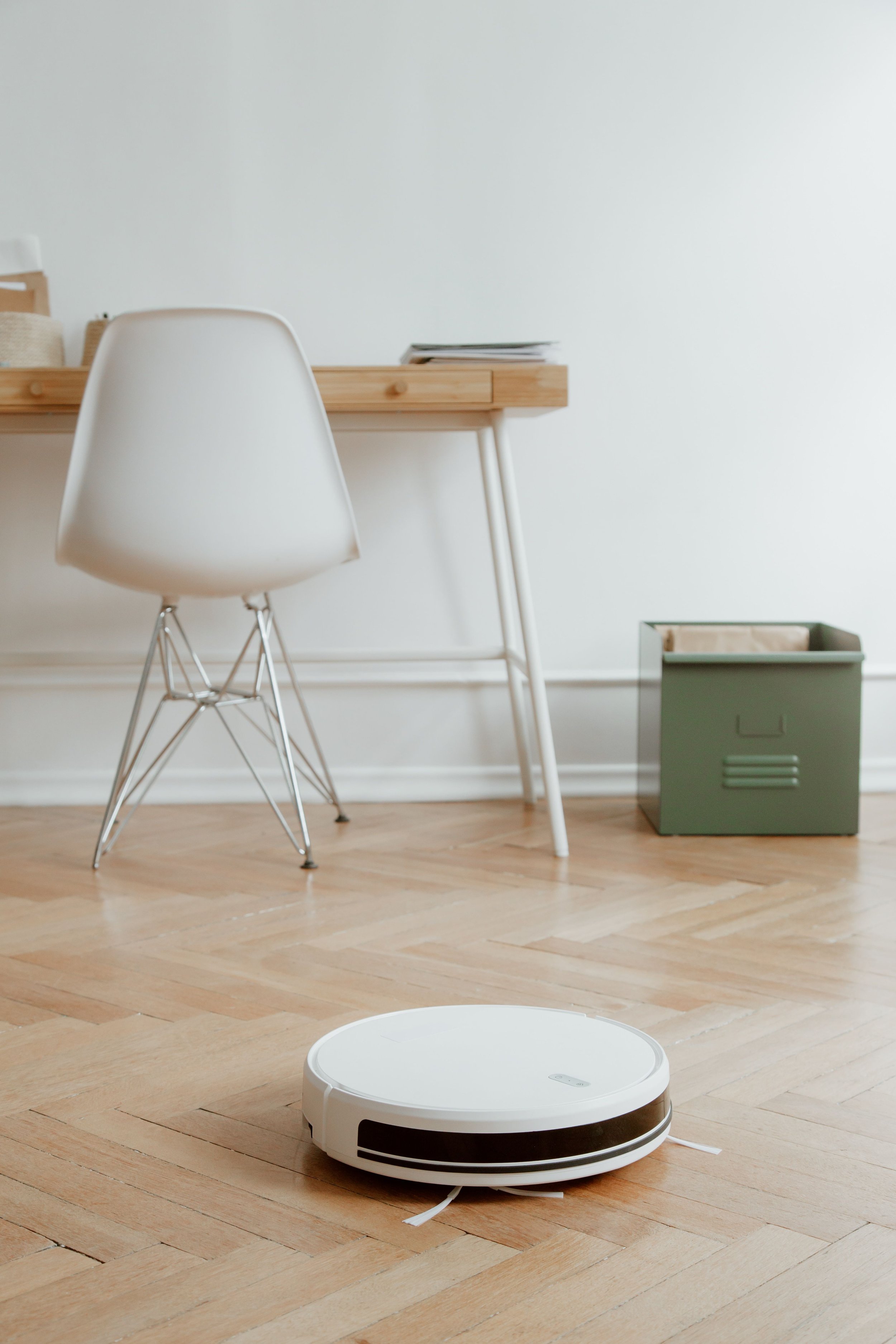 A robotic vacuum cleaner on a wooden floor in a minimalistic room with white walls, a wooden desk, a white chair, and a green storage box.