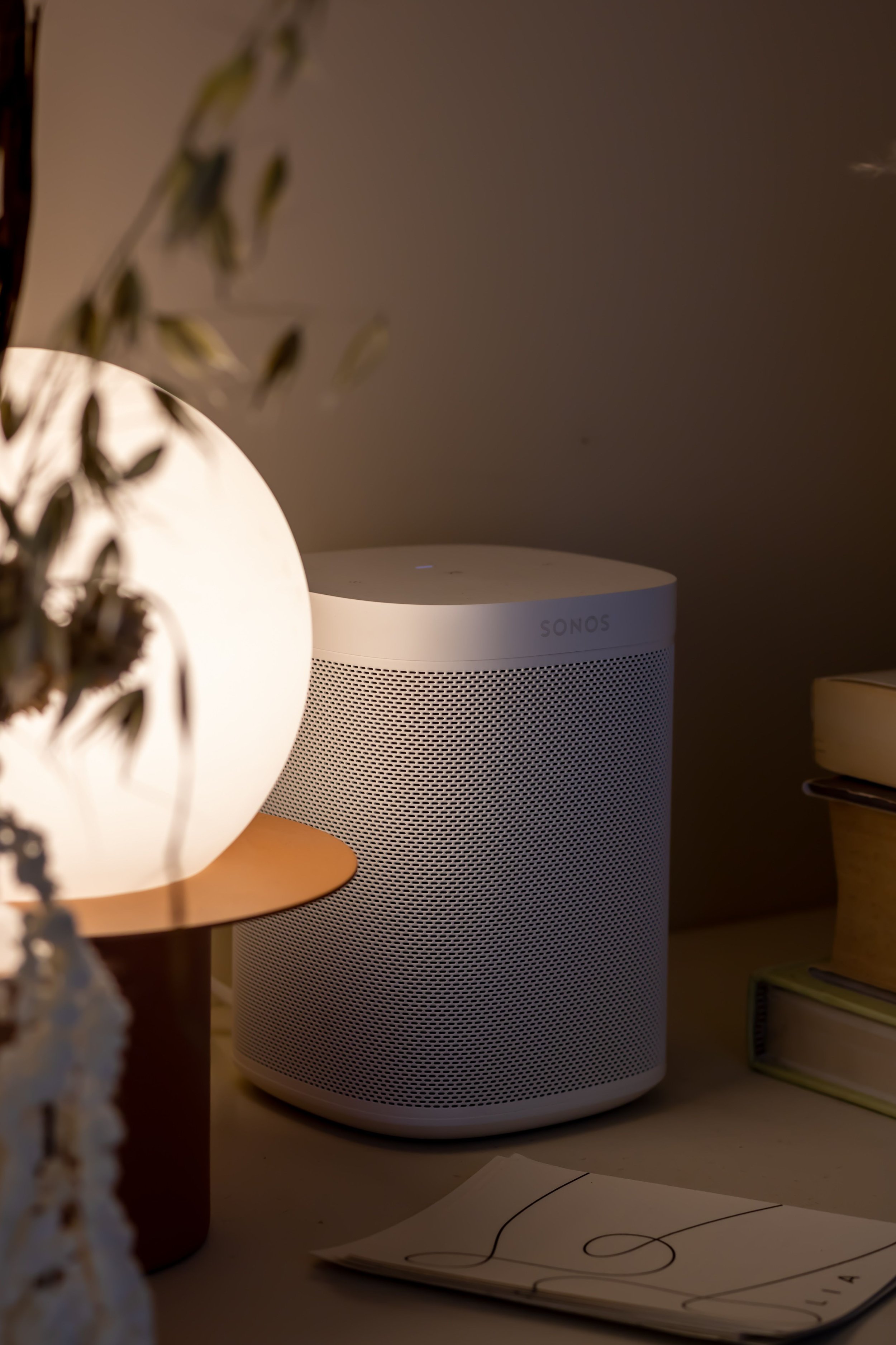 A white Sonos speaker placed on a flat surface in a cozy indoor setting, with a small book partly visible in the foreground and a lamp with a pear-shaped shade to the left.