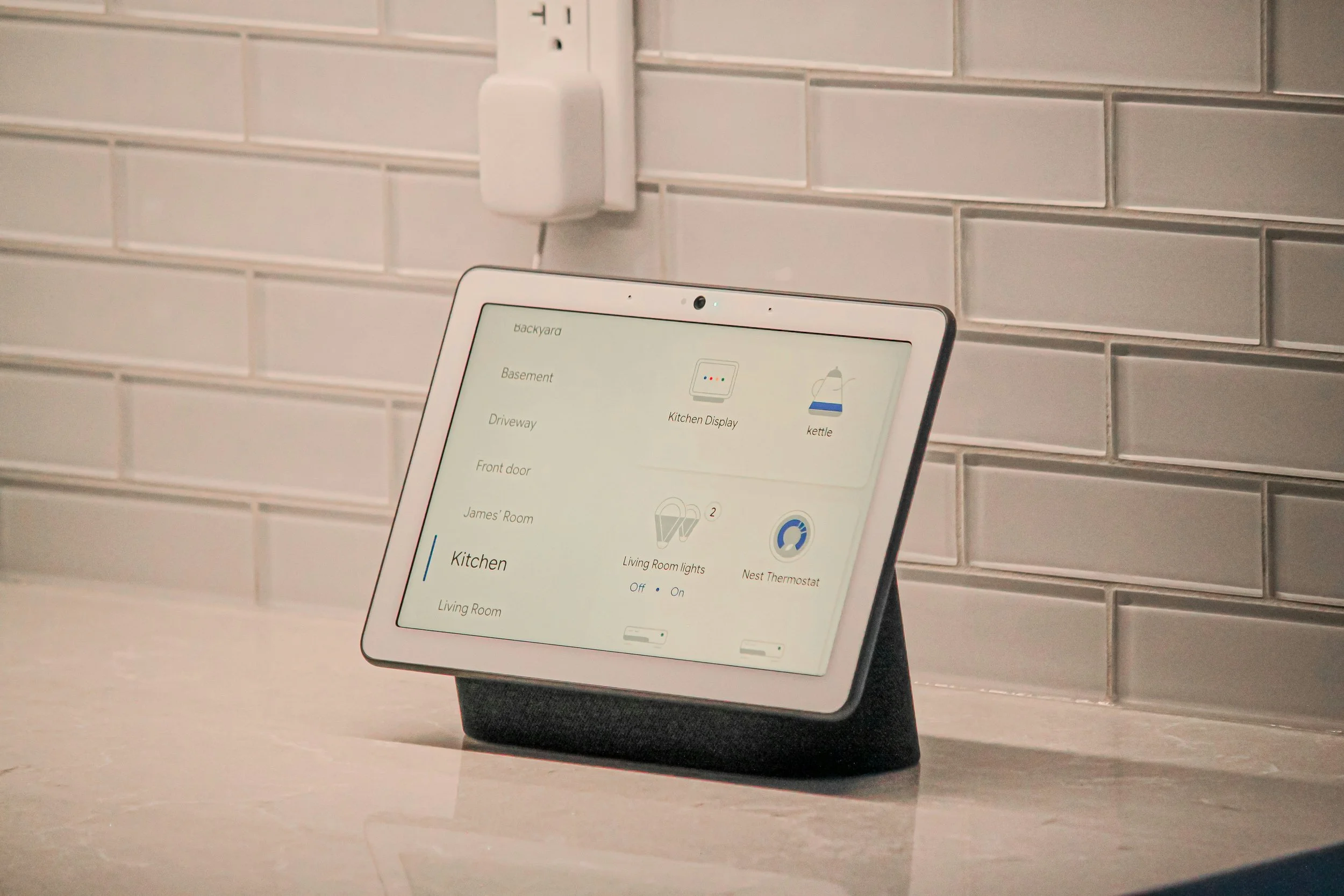 A tablet displaying a smart home control interface with options for different rooms and devices, resting on a countertop against a white tiled wall with an electrical outlet.