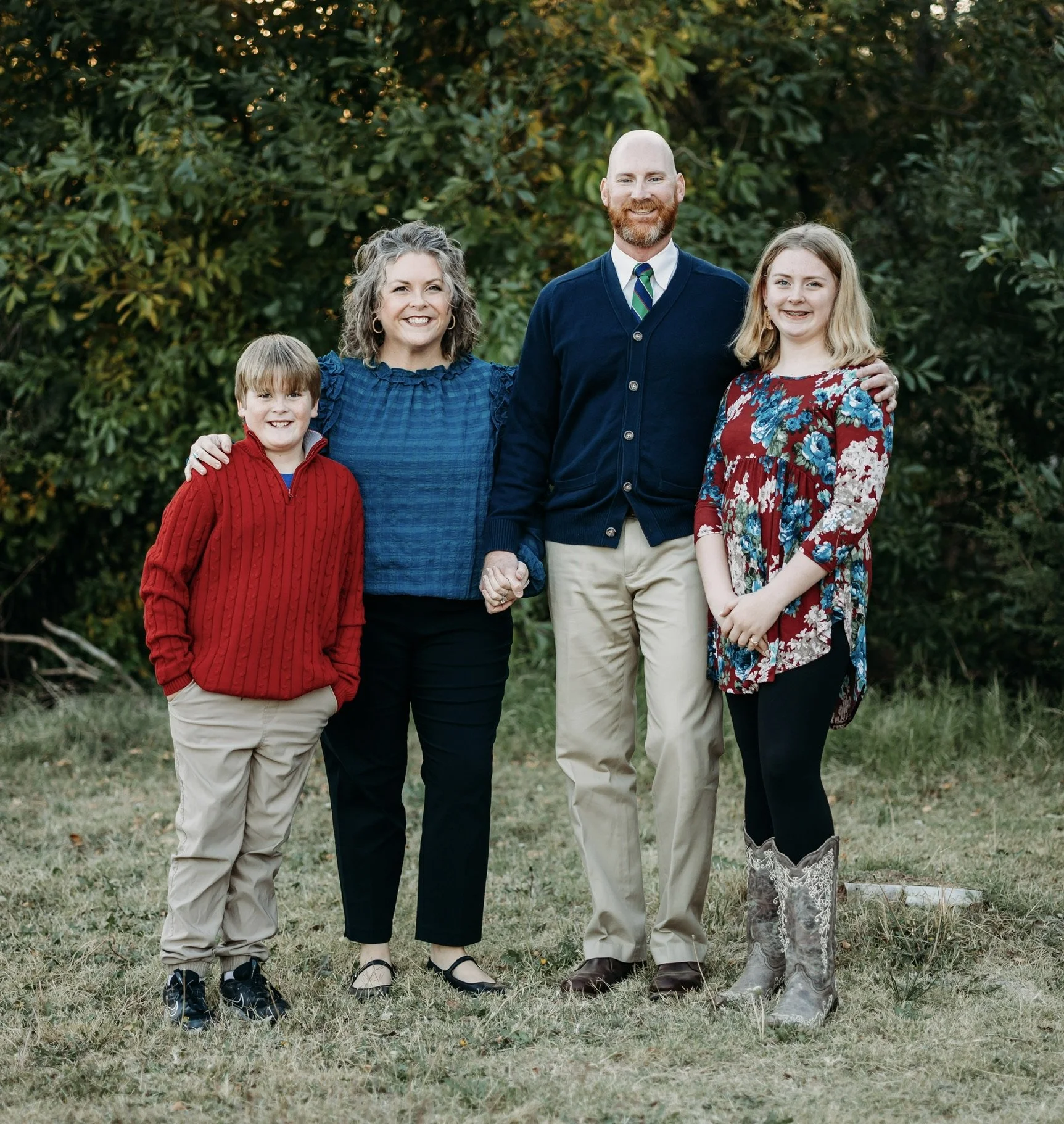 A family of five posing outdoors in front of green foliage, holding hands and smiling.