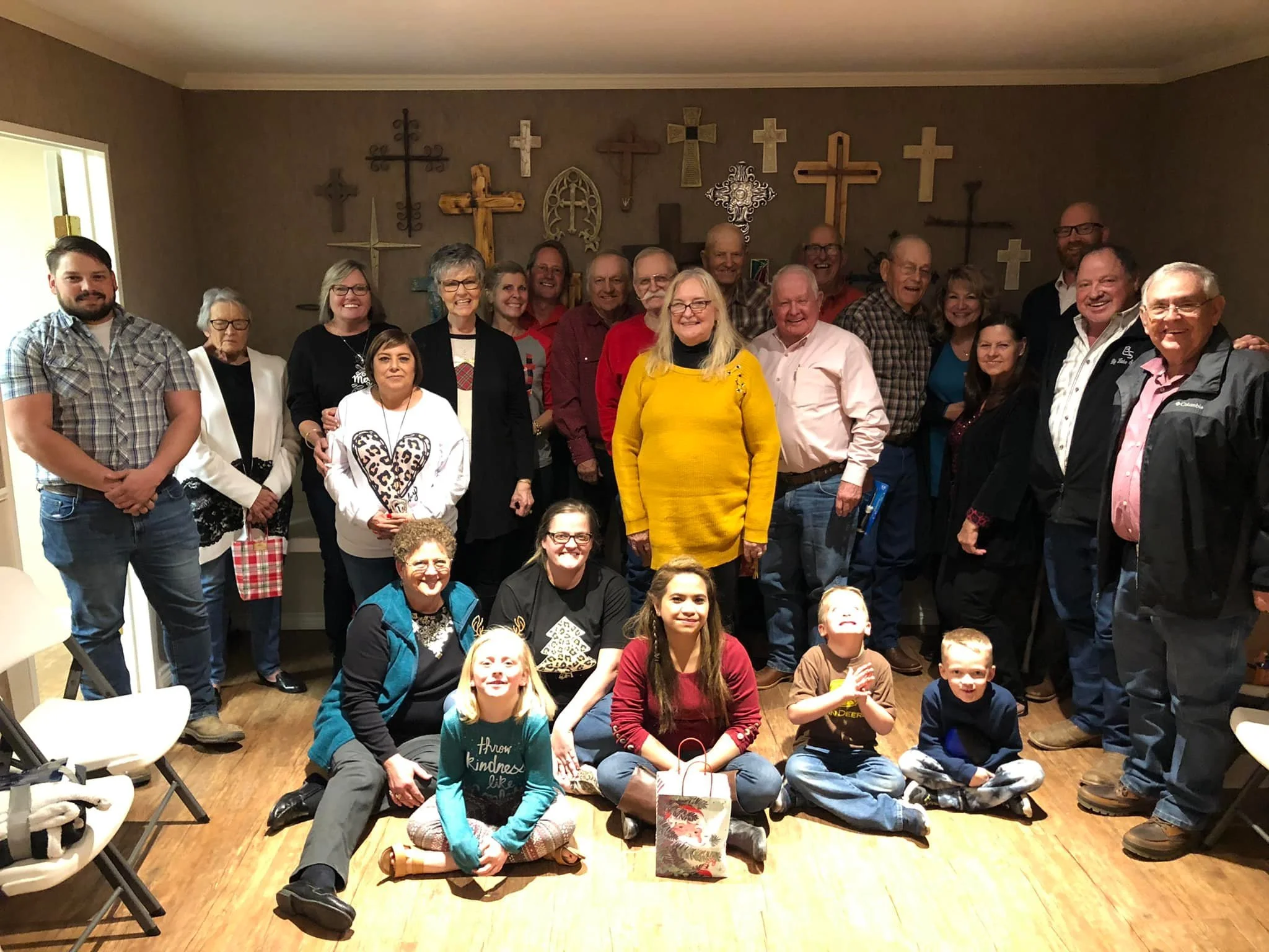 A large group of people, including men, women, and children, gathered indoors for a group photo in front of a wall decorated with various crosses. Some individuals are standing while others are sitting or kneeling on the floor, all smiling and looking at the camera.