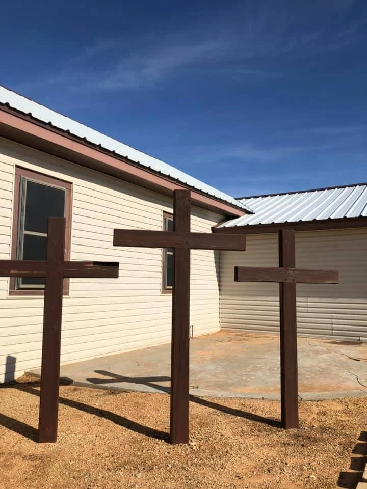 Three wooden crosses in front of a beige building with a metal roof, under a clear blue sky.