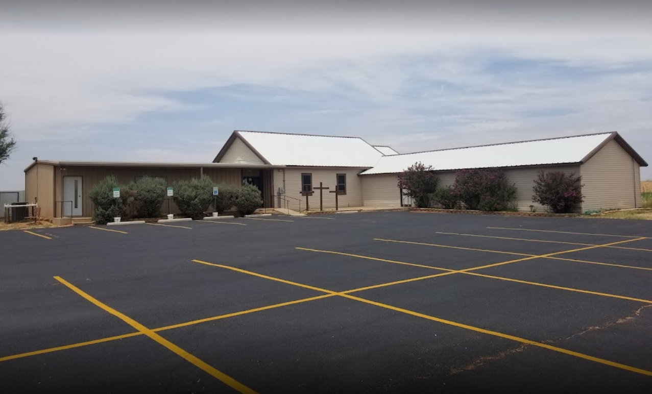 Empty parking lot in front of a beige church building with a cross display, bushes, and a partly cloudy sky.