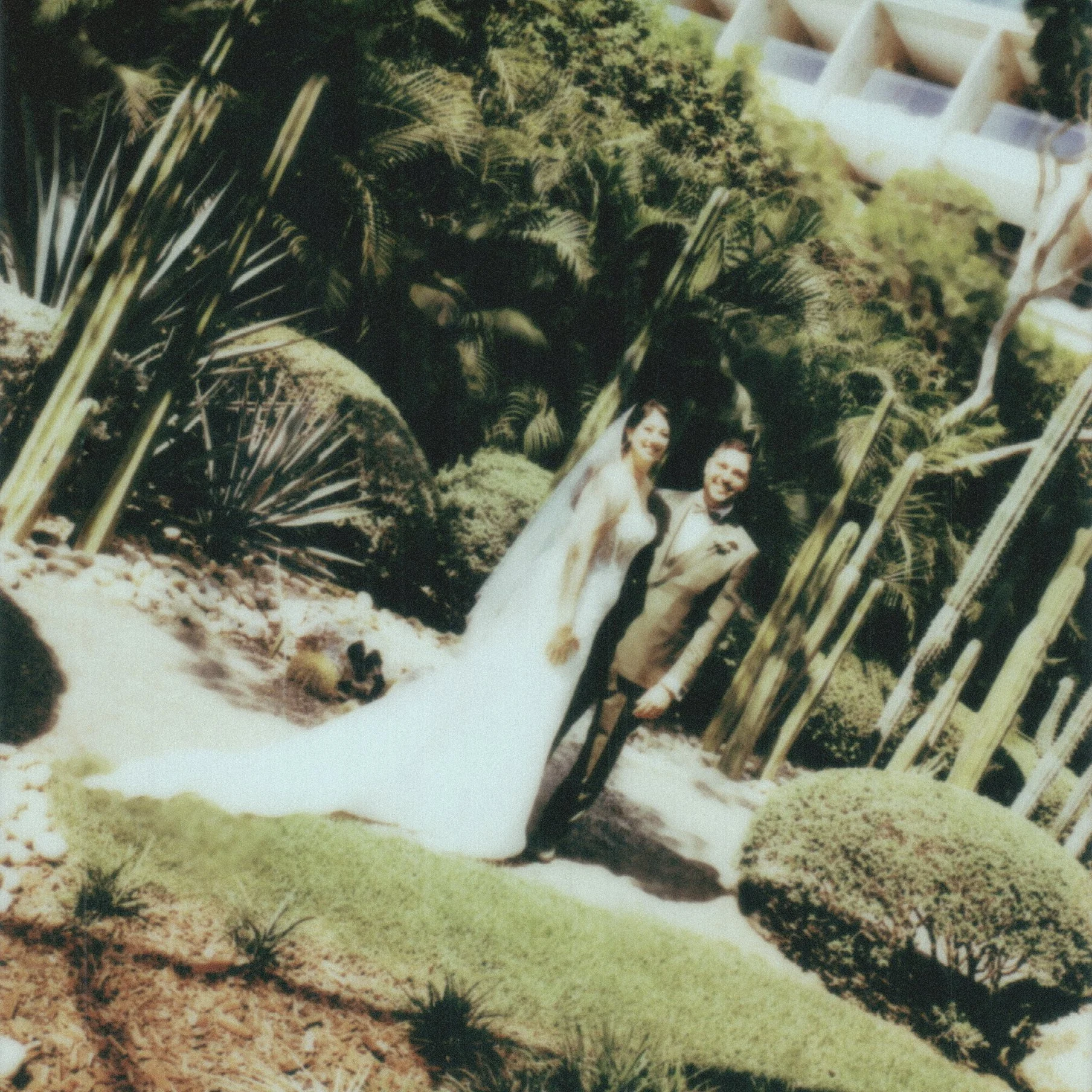 A miniature model of a garden scene with small trees, rocks, and a white fence, featuring two small happy people figurines, one woman and one man, dressed in white.
