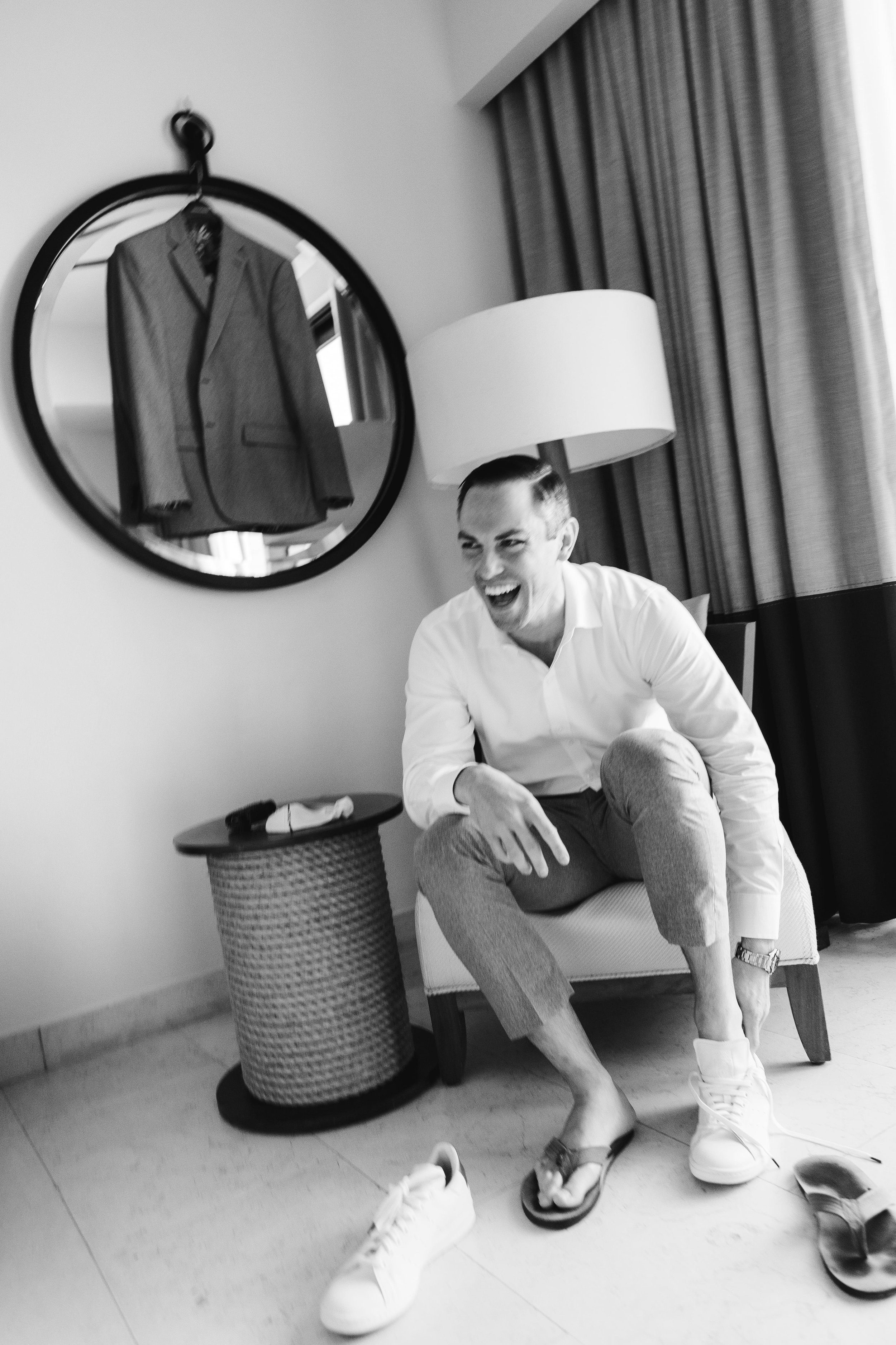 A smiling man sitting on a chair in a room with a reflective mirror showing a suit hanging, a lamp, and curtains behind him.