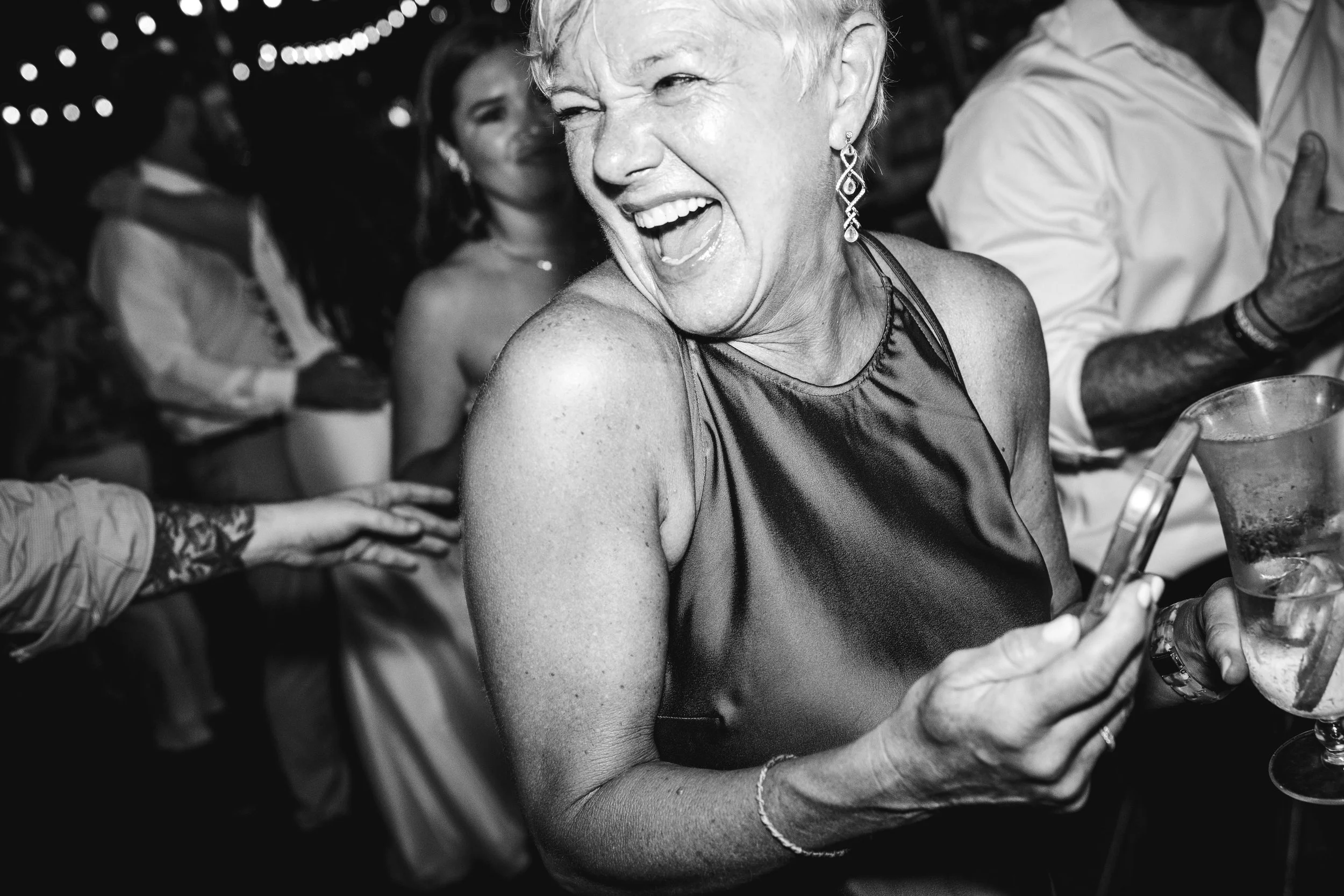 A black and white photo of a woman with short, light-colored hair, laughing joyfully at a social event, holding a drink in her hand. She is wearing earrings and a sleeveless top, with others blurred in the background.
