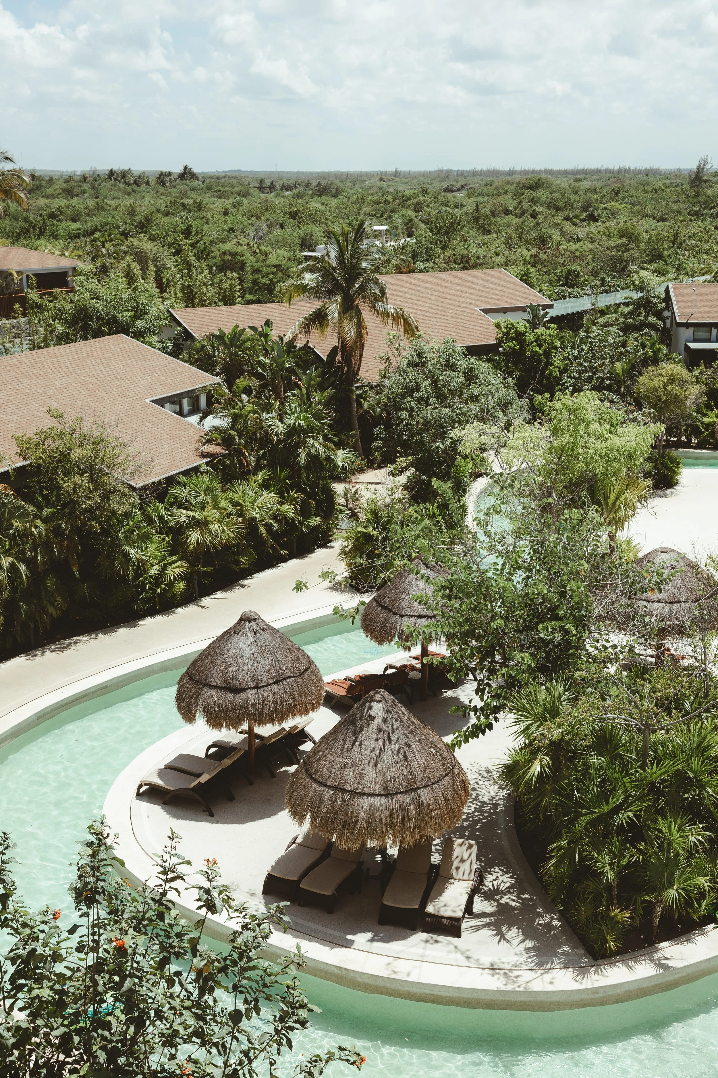 Pool area with sun loungers and thatched umbrellas, surrounded by lush tropical greenery, residential rooftops, and a distant forest under a cloudy sky.